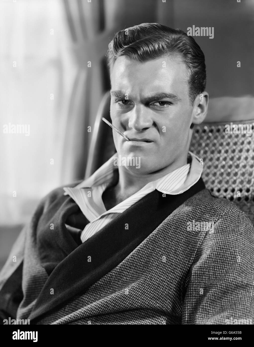1930s 1940s UNHAPPY MAN SITTING HOLDING THERMOMETER IN MOUTH WEARING ROBE PAJAMAS Stock Photo