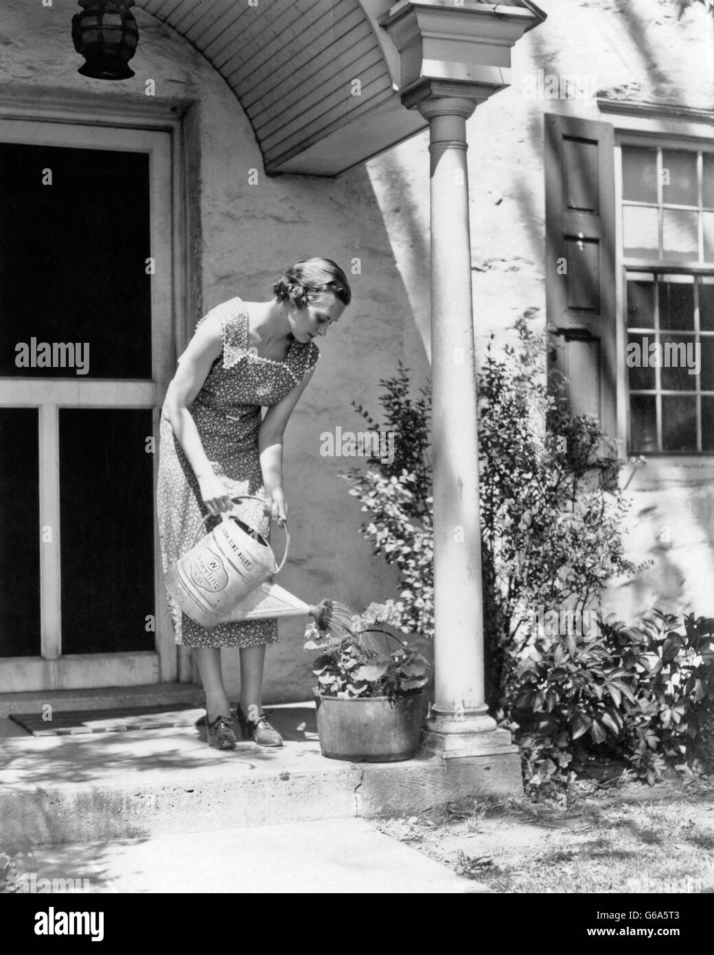 1930s WOMAN WEARING PRINT DRESS WATERING POTTED PLANT WITH WATERING CAN ON FRONT STOOP OF HOUSE Stock Photo