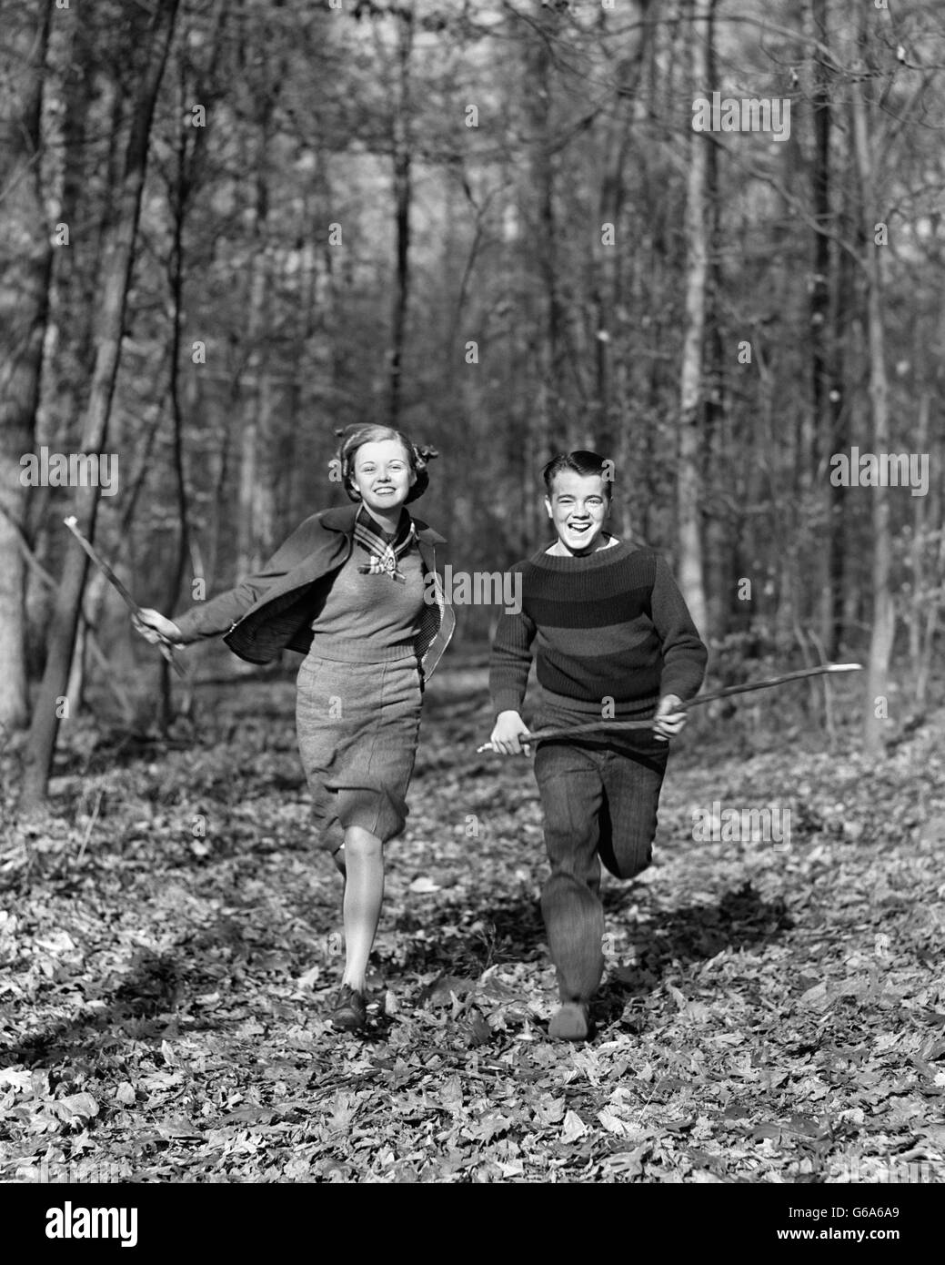 1940s SMILING TEENAGE COUPLE BOY GIRL RUNNING IN AUTUMN WOODS LANDSCAPE Stock Photo