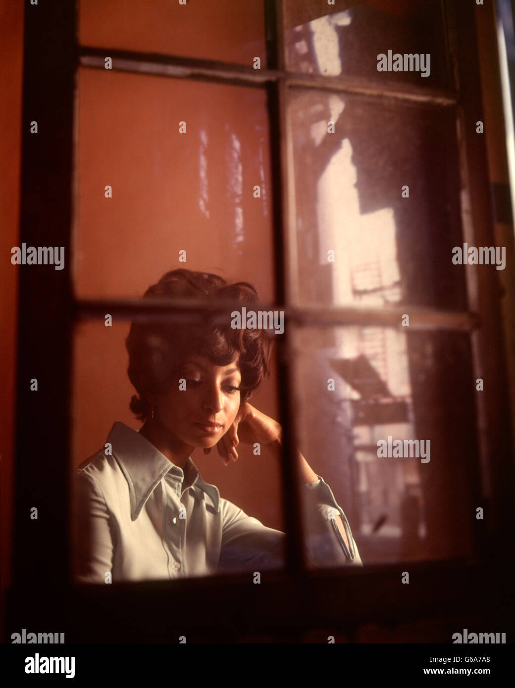 1970s SAD THOUGHTFUL SERIOUS TROUBLED AFRICAN AMERICAN WOMAN SITTING BEHIND WINDOW REFLECTION OF BUILDING FIRE ESCAPE Stock Photo