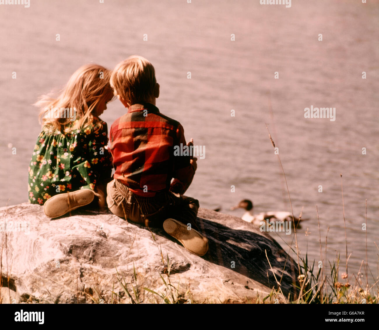 1970s TWO CHILDREN BOY GIRL SITTING TOGETHER ON A ROCK OVERLOOKING A LAKE FEEDING DUCKS Stock Photo