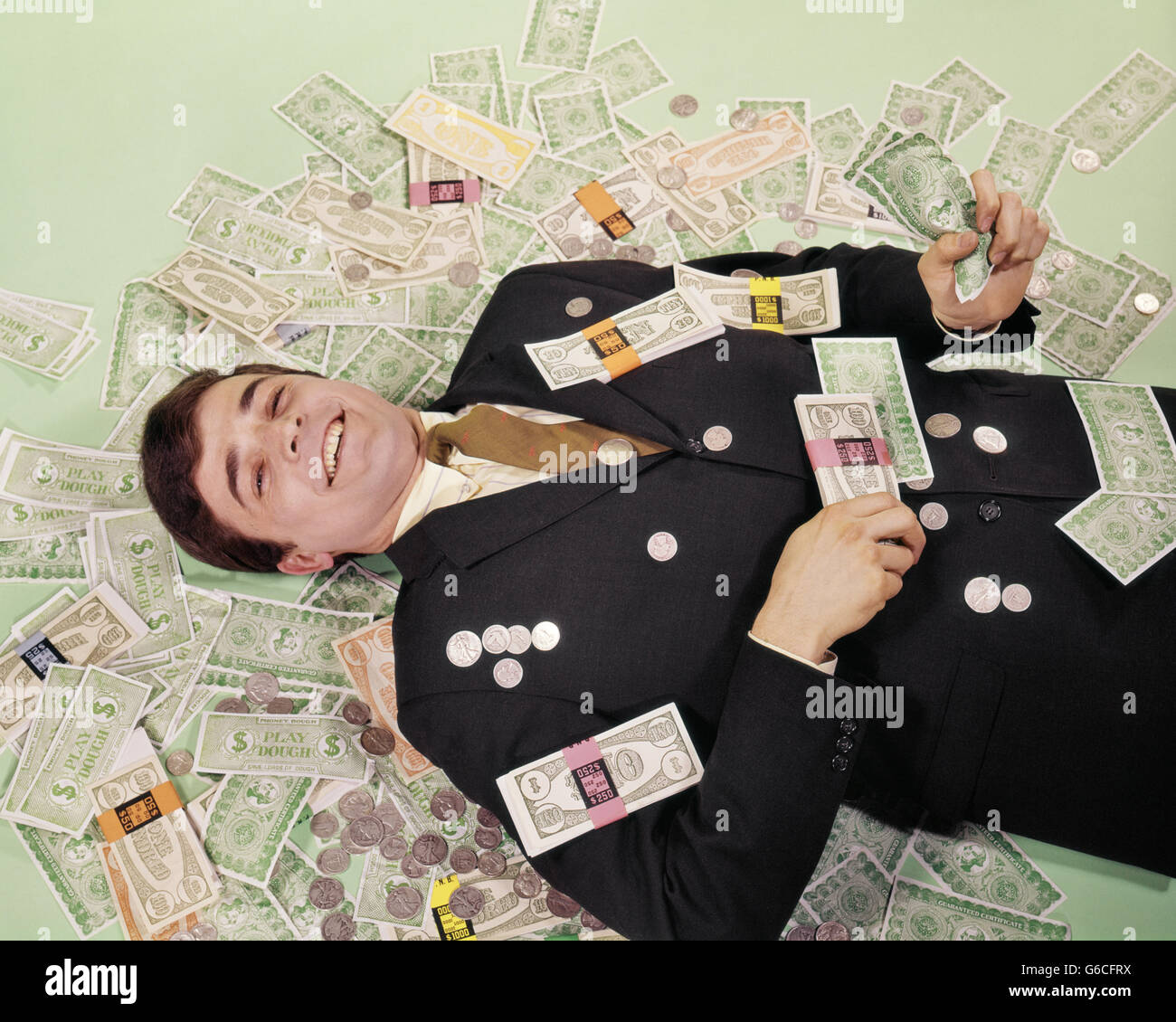 1960s SMILING MAN LYING ON FLOOR COVERED WITH FAKE MONEY Stock Photo