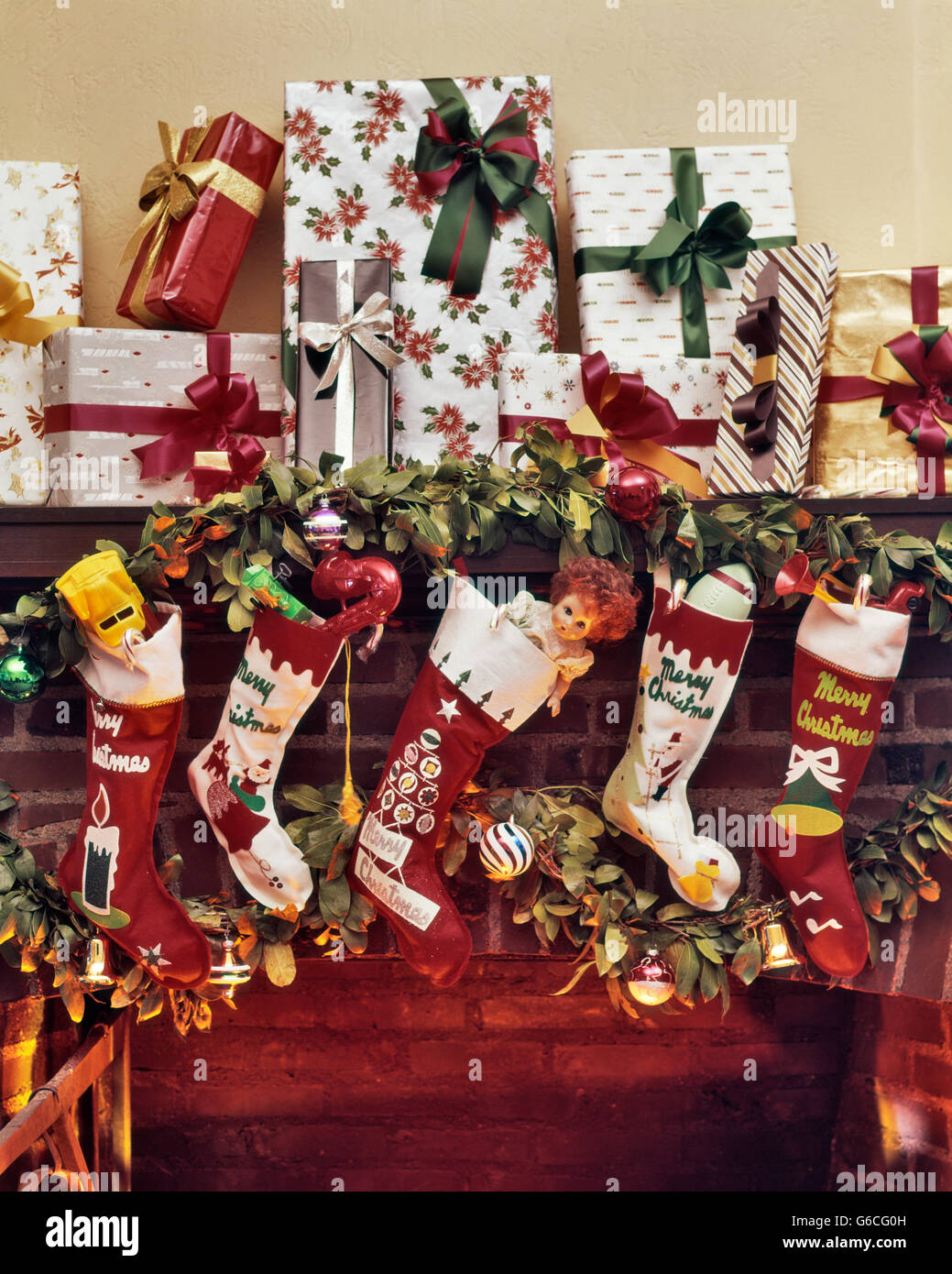 1960s FIVE CHRISTMAS STOCKINGS HANGING ON FIREPLACE MANTLE LOTS OF WRAPPED PRESENTS STACKED ABOVE Stock Photo
