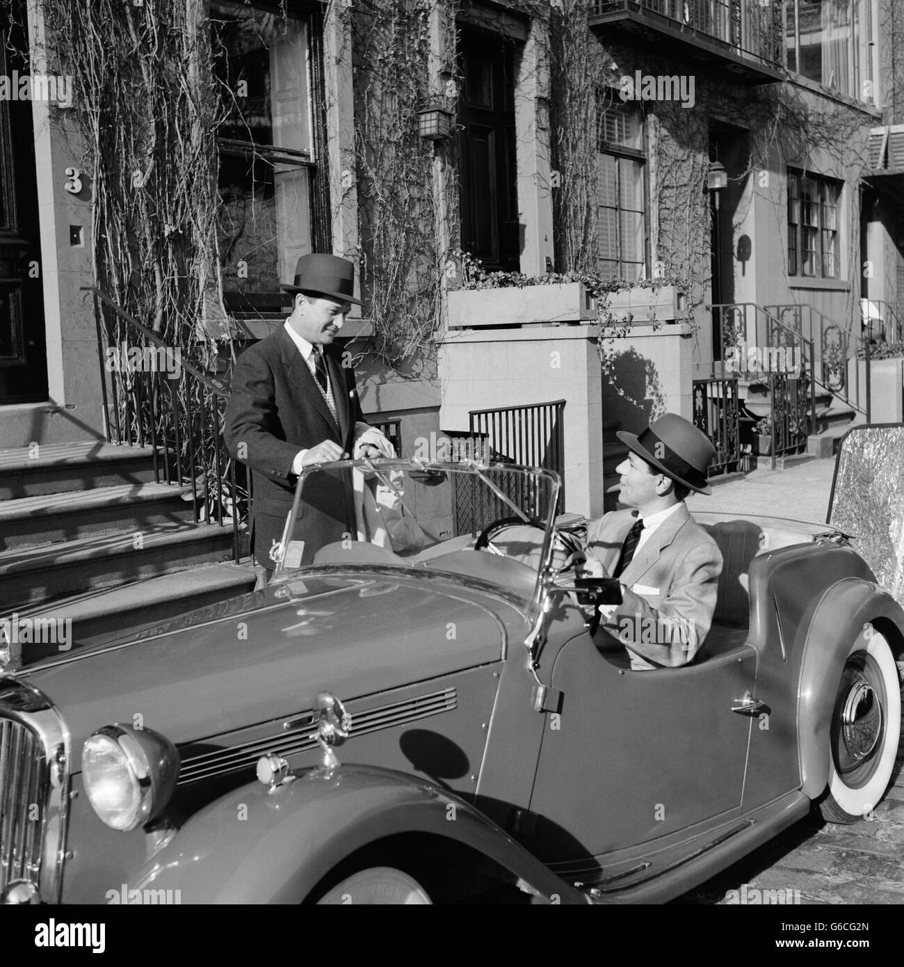 1950s 1960s TWO BUSINESSMEN TALKING ON STREET ONE SITTING IN CONVERTIBLE TWO SEAT ENGLISH SPORTS CAR Stock Photo