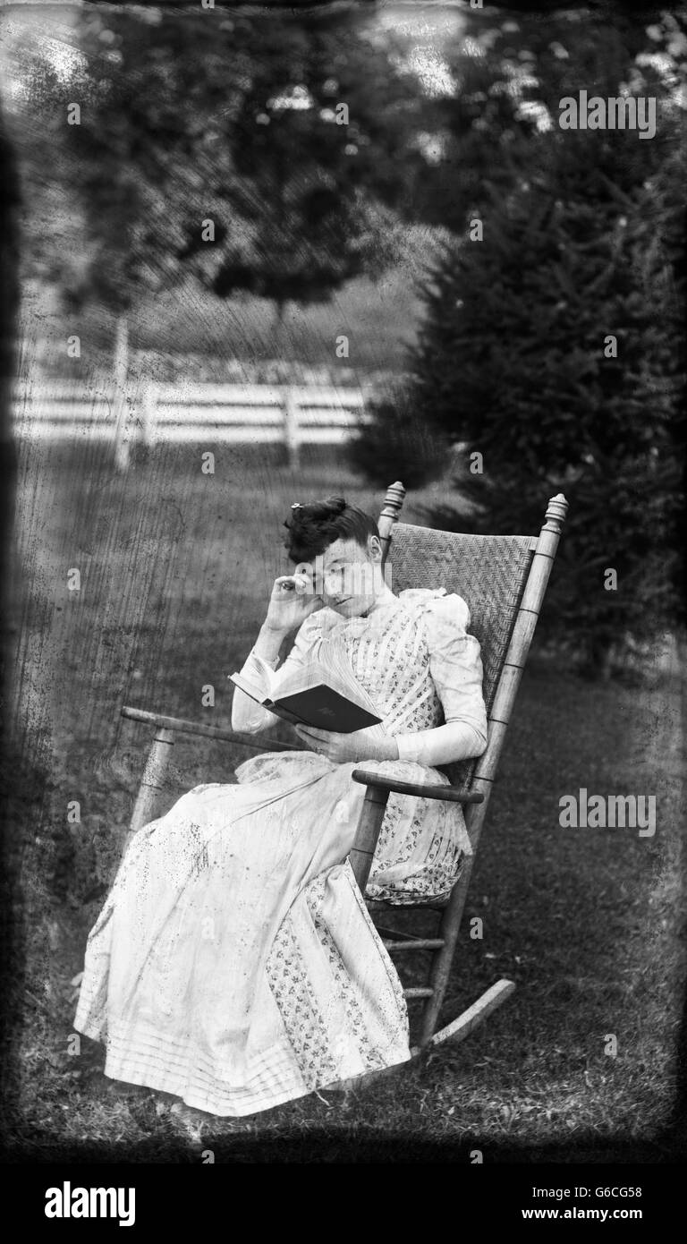1890s WOMAN SITTING IN ROCKING CHAIR IN BACKYARD READING BOOK Stock Photo