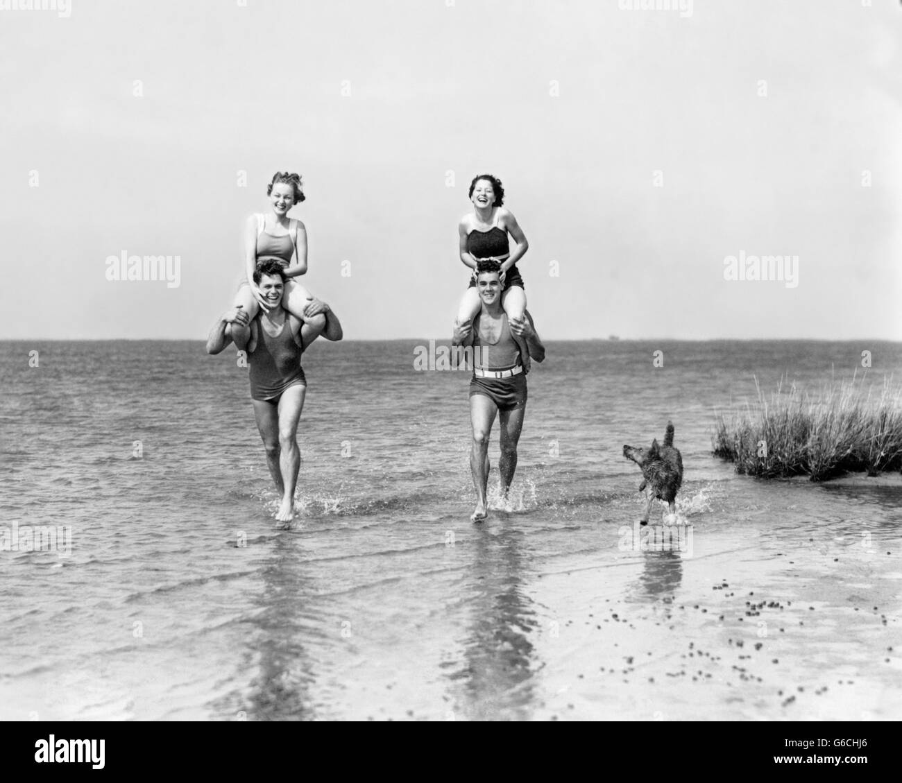 1930s TWO COUPLES RUNNING IN OCEAN WEARING BATHING SUITS GIRLS SITTING ON BOYS SHOULDERS AND DOG RUNNING ALONG SIDE Stock Photo