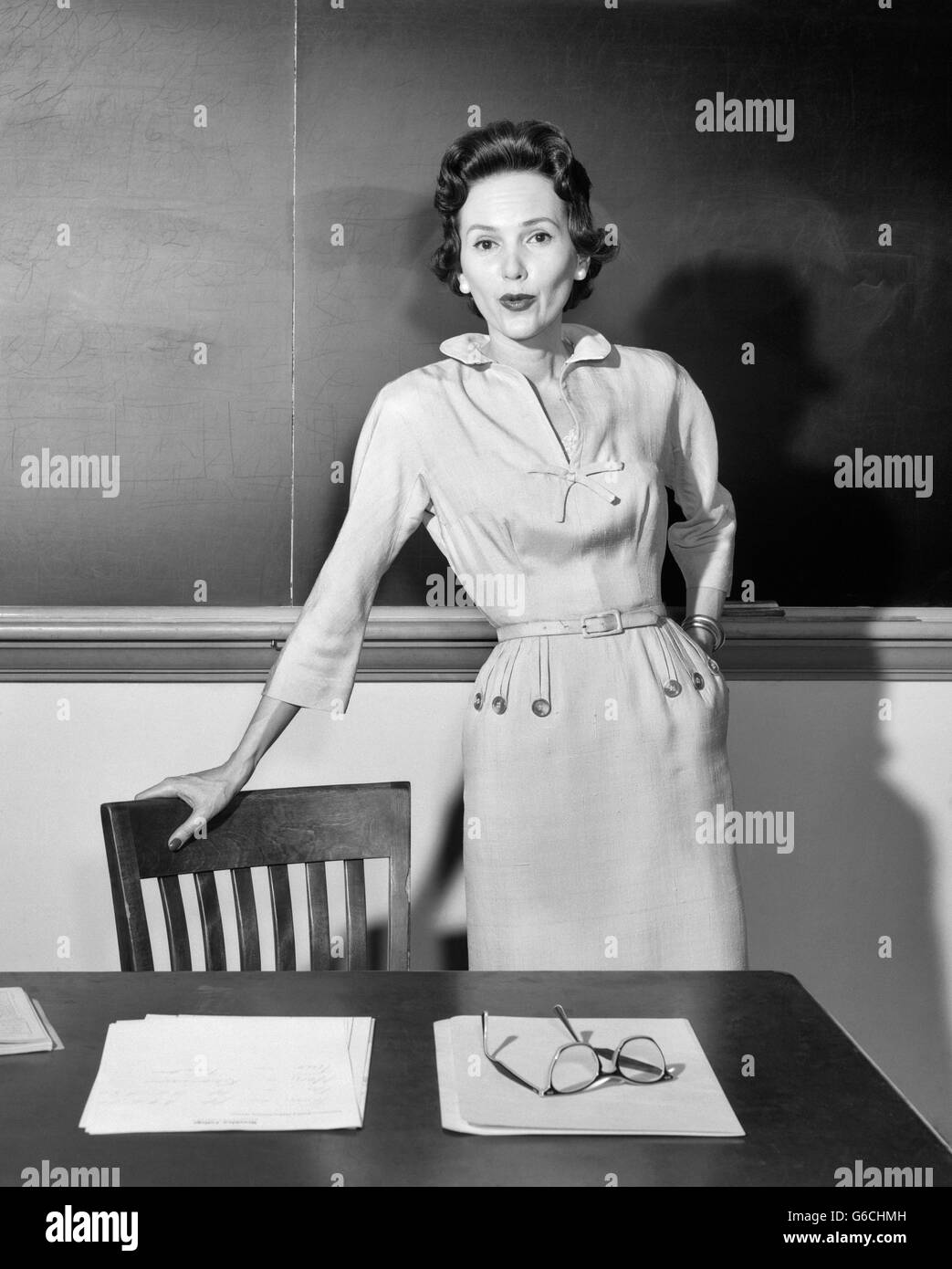 1950s WOMAN SCHOOL TEACHER LOOKING AT CAMERA STANDING IN FRONT OF CHALKBOARD IN CLASSROOM LEANING ON CHAIR BY DESK Stock Photo