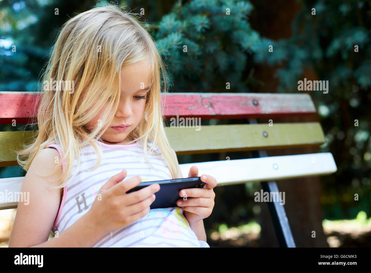 Beautiful Caucasian blond girl sitting on old park bench playing on smart phone Stock Photo
