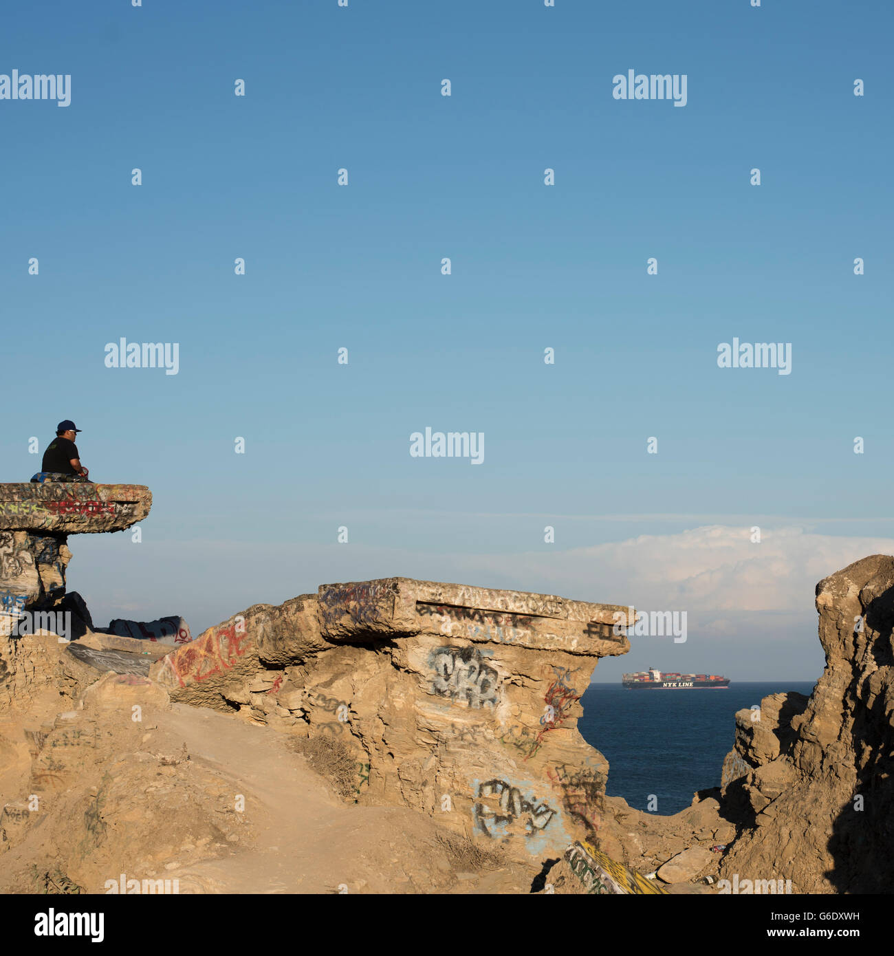 A man sits on graffiti covered slab of concrete as a distant ship heads to sea at Sunken City in San Pedro, Calif., on Aug. 21, 2014. Stock Photo