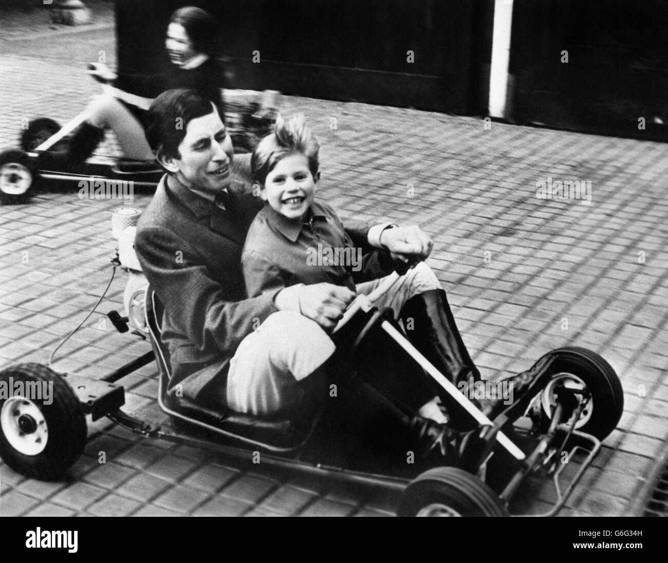 Prince Charles gives his five-year-old brother Prince Edward a ride on a go-kart in the grounds of Windsor Castle. Princess Anne is driving her own kart in the background. Stock Photo