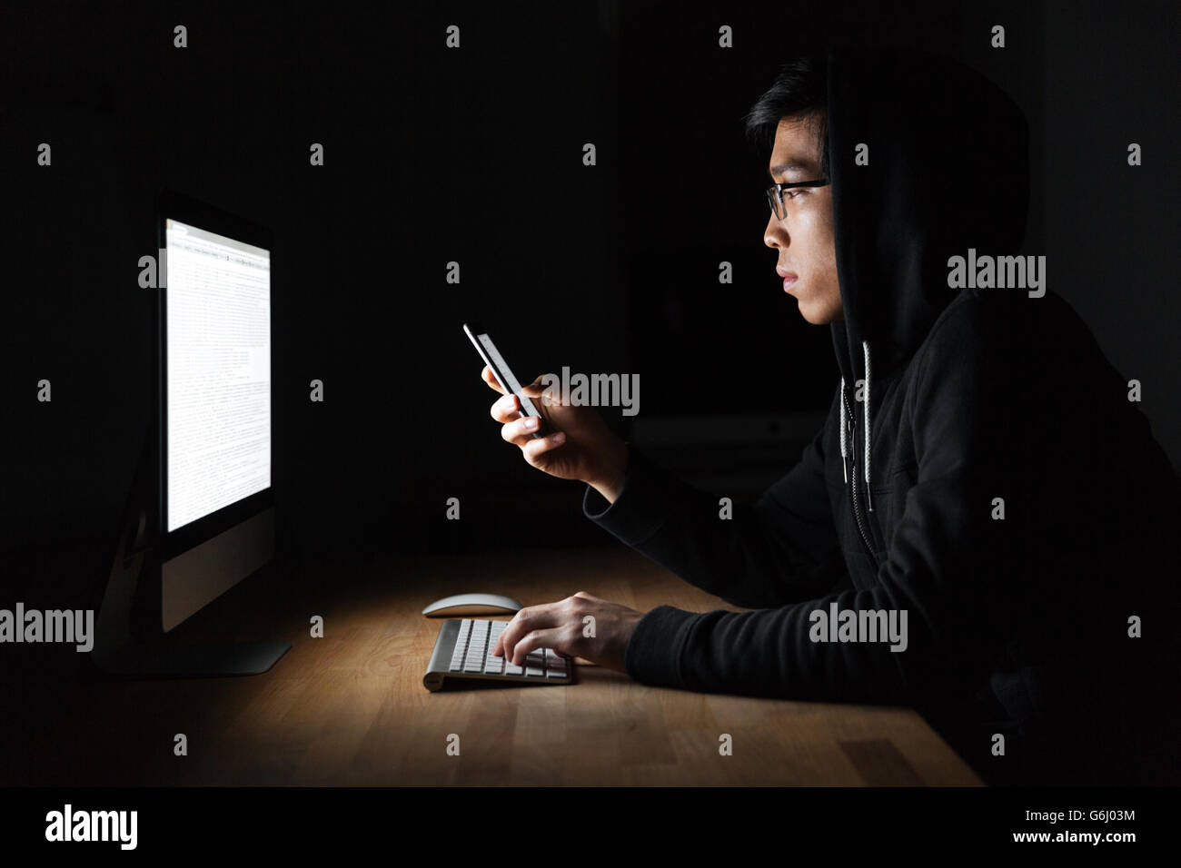Serious young hacker using laptop and mobile phone in dark room Stock Photo