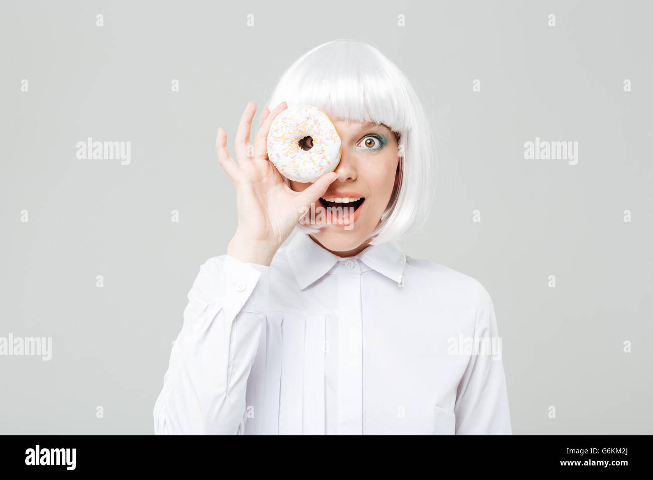Cheerful young woman in blonde wig covered her eye with donut over white background Stock Photo