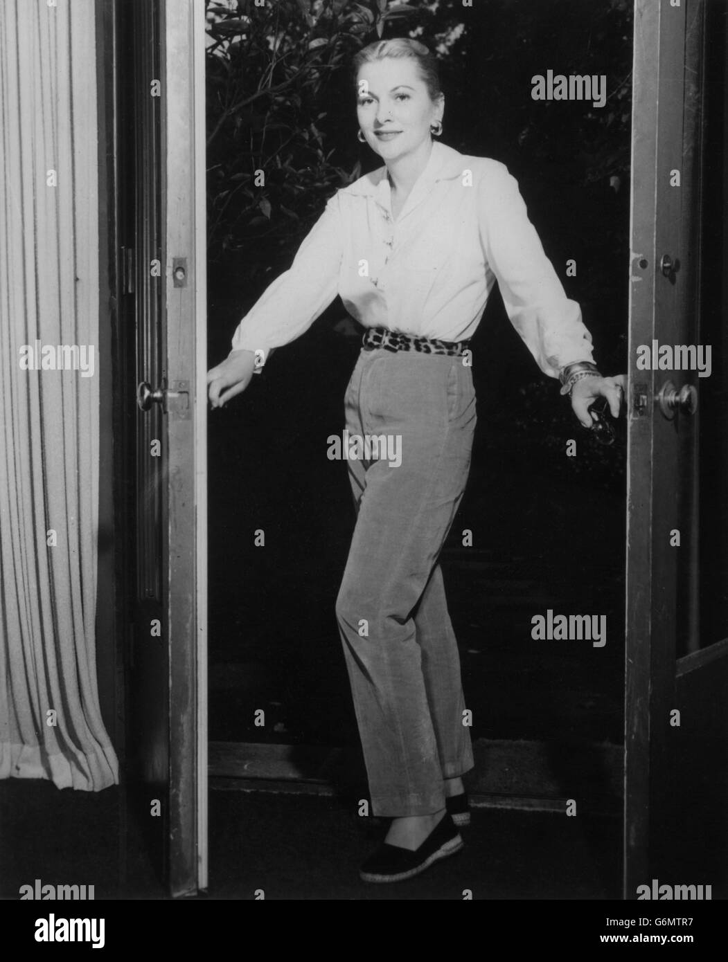 Actress Joan Fontaine, who is now starring in MGM's 'Until They Sail', likes to wear comfortable casual clothing around the house. She is seen here in mustard velveteen trousers teamed teamed with a white Italian silk blouse having hand-wrought gold buttons. A leopard belt and black espadrilles complete the costume. Stock Photo