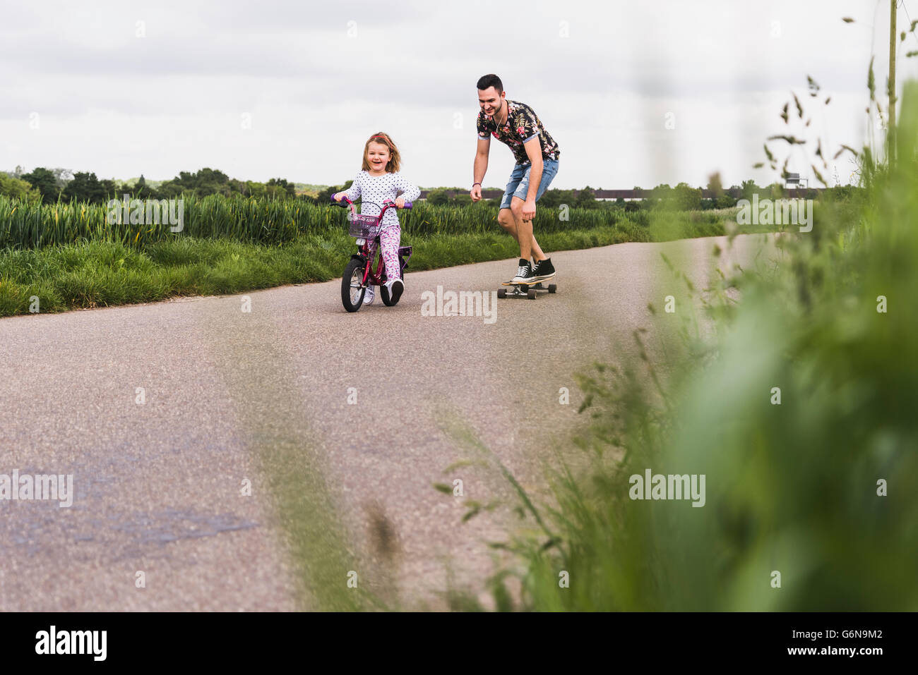 Father on skateboard accompanying daughter on bicycle Stock Photo