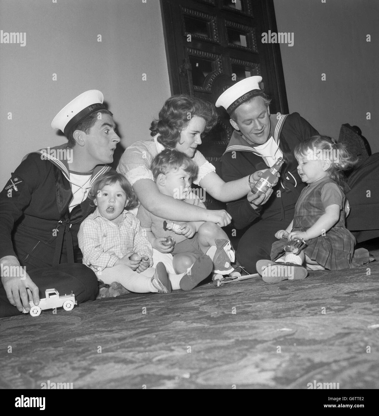 During the interval of comedy 'Rock-a-Bye Sailor', actors Ian MacNaughton (l) and Ian Curry visit a special nursery to entertain children and the nannie, 20-year-old Annette Miller, at the Phoenix Theatre in Charing Cross Road, London. The youngsters are (from l-r) Katie McKenzie, Simon Bentley and Janie Karen Mills, all two years old. The nursery has been set up to enable mothers to attend the matinees performance of the comedy show. Stock Photo