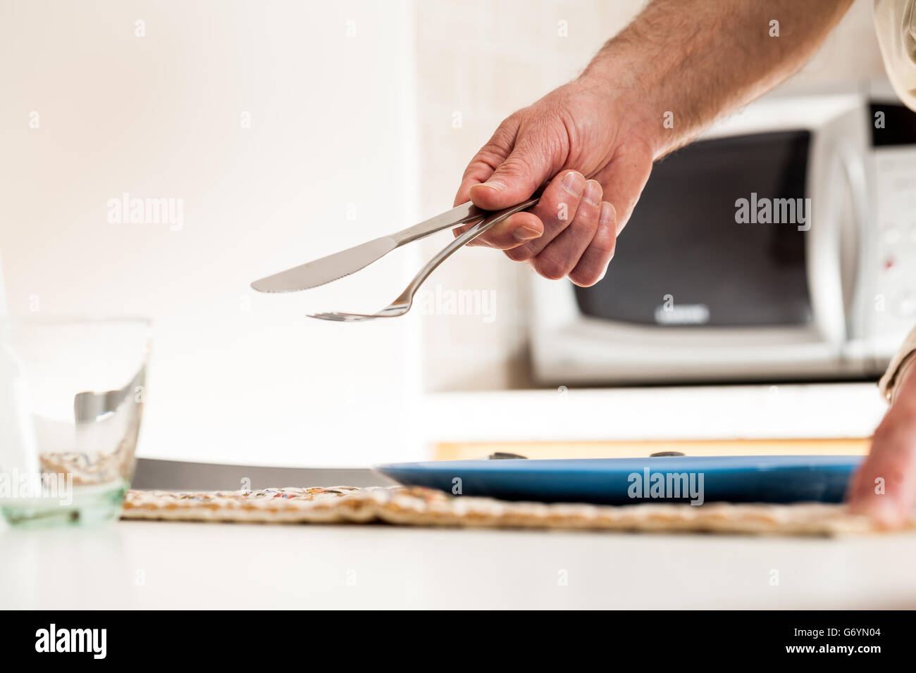 Close up of hand holding knife and fork at table with glass and plate underneath and microwave oven in background Stock Photo