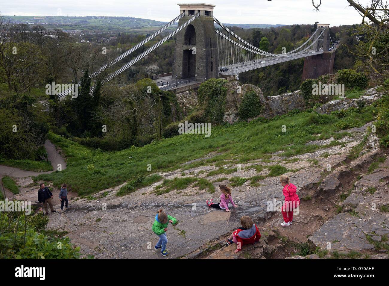 Children slide down a rock seam, worn smooth by repeated use, on the side of a hill, overlooking Clifton Suspension Bridge, Bristol. Stock Photo