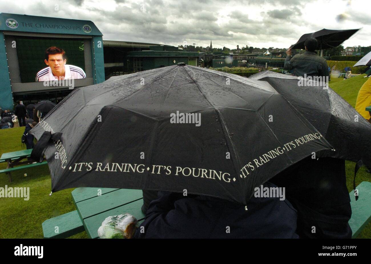 Tennis fans take cover under umbrellas as they watch repeats of matches on the big screen at The Lawn Tennis Championships in Wimbledon, London today, where rain and gales delayed the start of play. 24/06/04: Players and spectators at Wimbledon were praying for good weather in the hope that heavy rain would not destroy another day of tennis. For only the eighth time in the last 30 years the third day's play at this year's championships was a complete wash-out, as persistent downpours and strong winds hit SW19. Stock Photo