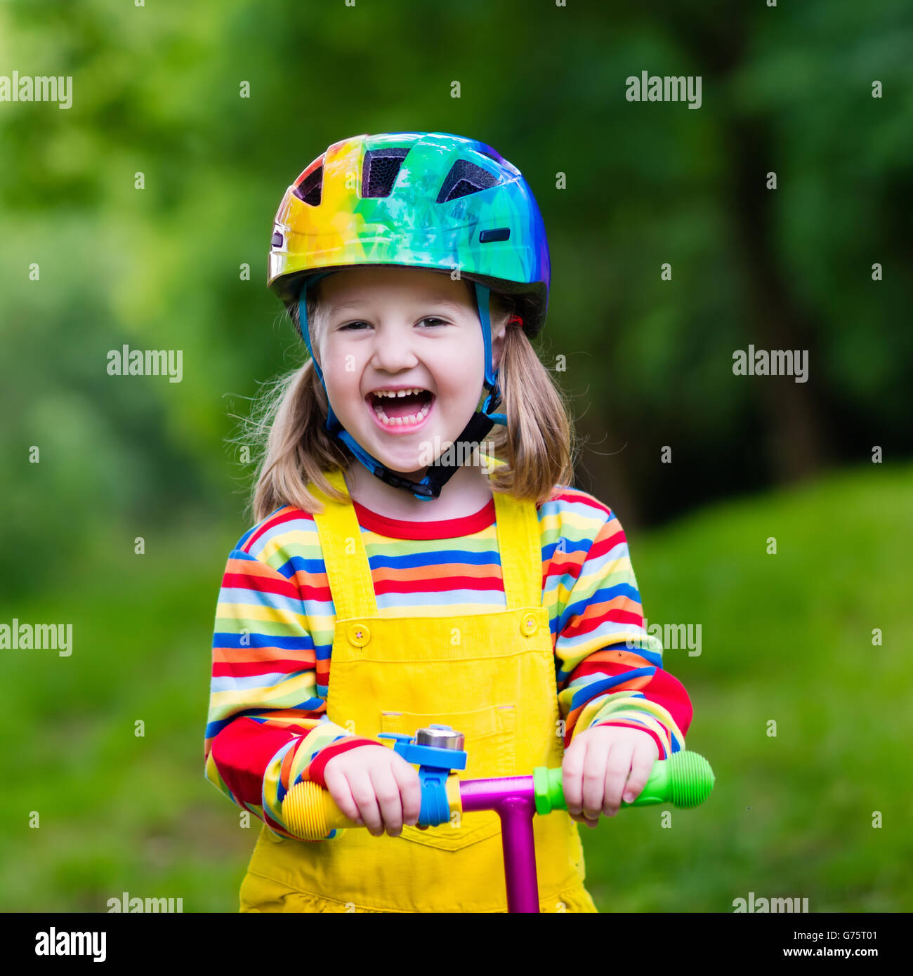 Little child learning to ride a scooter in a city park on sunny summer day. Cute preschooler girl in safety helmet riding roller Stock Photo