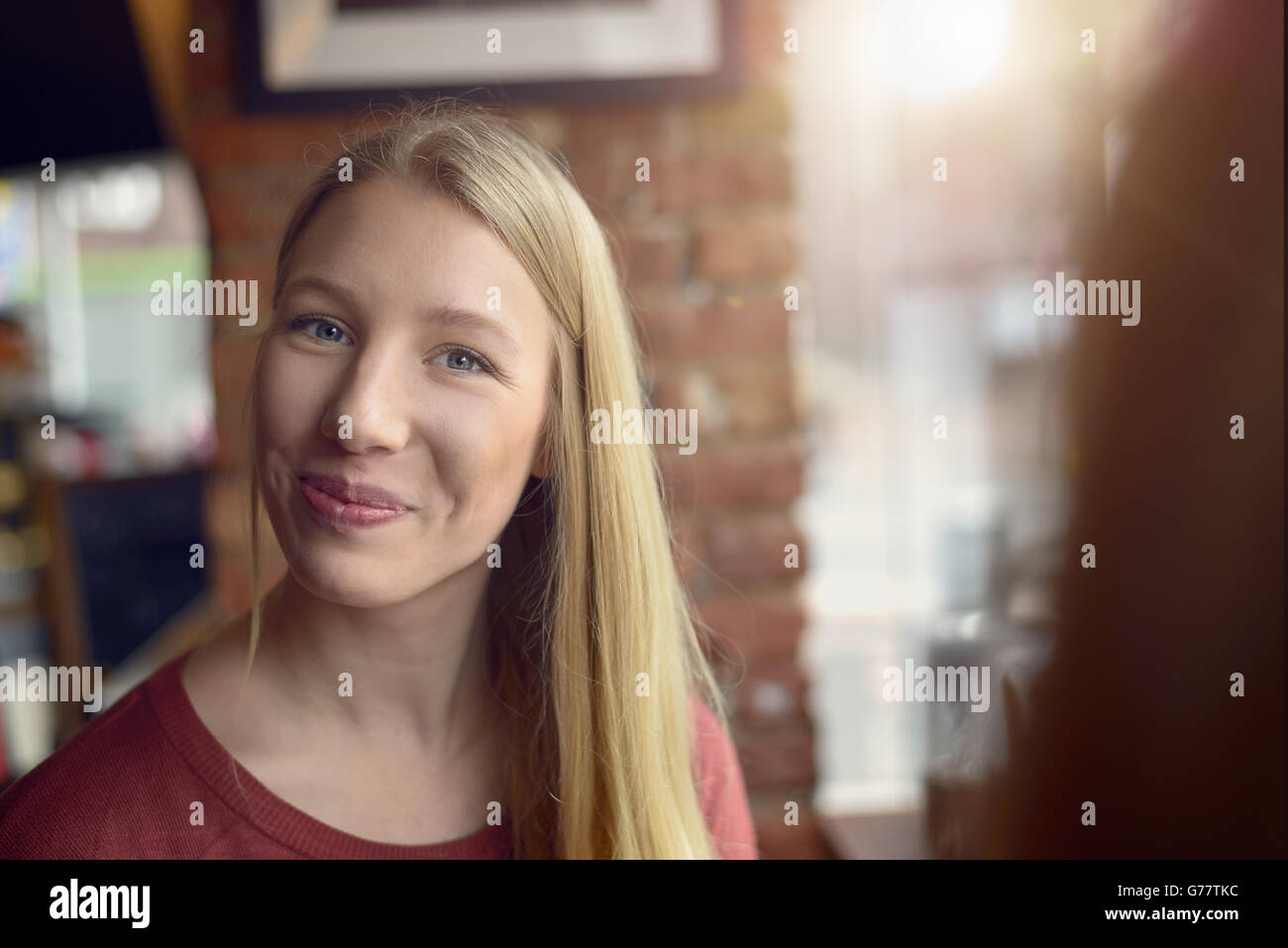 Smiling pretty young woman with long blond hair and an amused look gazing at the camera with a friendly smile inside a restauran Stock Photo