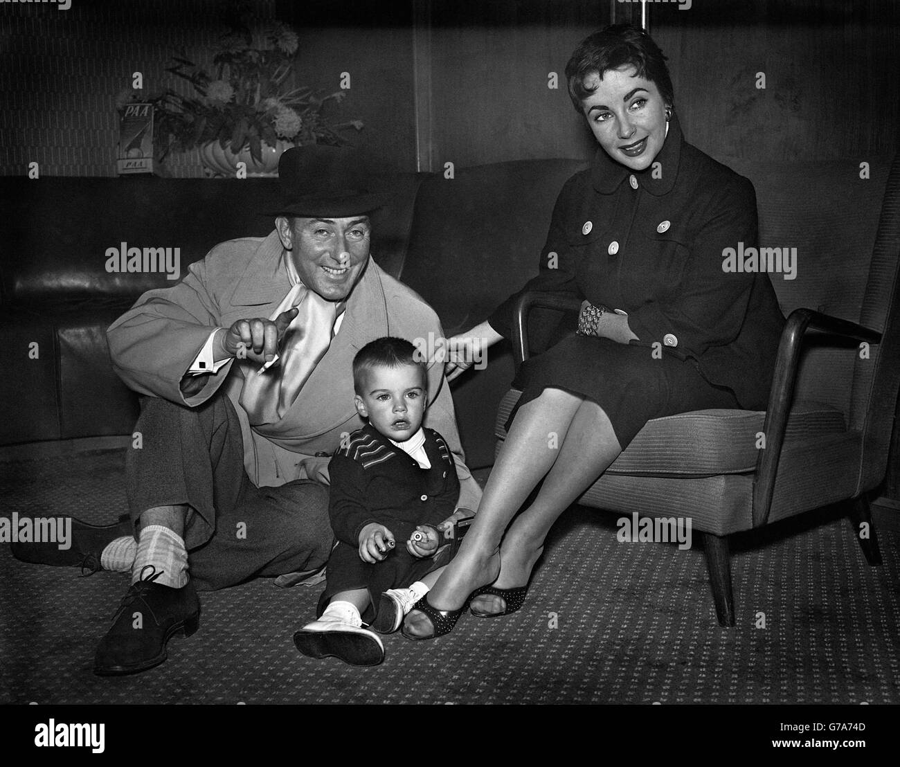 Actor Michael Wilding gets down on the floor to play with his two-year-old son Michael, Junior, as he waits with his wife, Elizabeth Taylor, at London Airport. Stock Photo