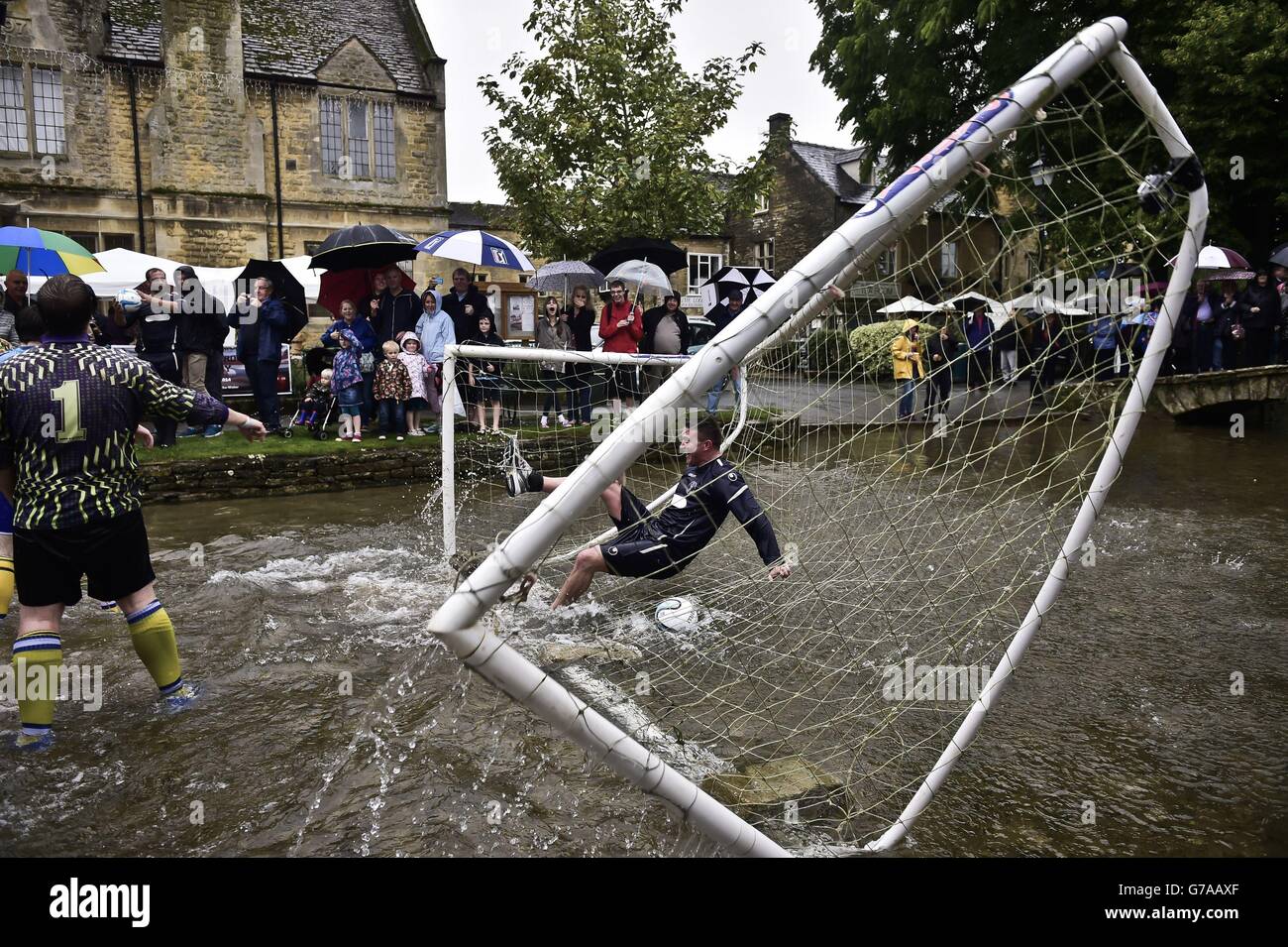 A Bourton Rovers 1st XI player crashes into the goal as Bourton Rovers 1st XI (dark blue) and Bourton Rovers 2nd XI (light blue) battle it out in the annual Football In The River match at Bourton-in-the-Water, Gloucestershire, where a game of two 15 minute halves of football are played in the River Windrush on the high street in the village. Stock Photo