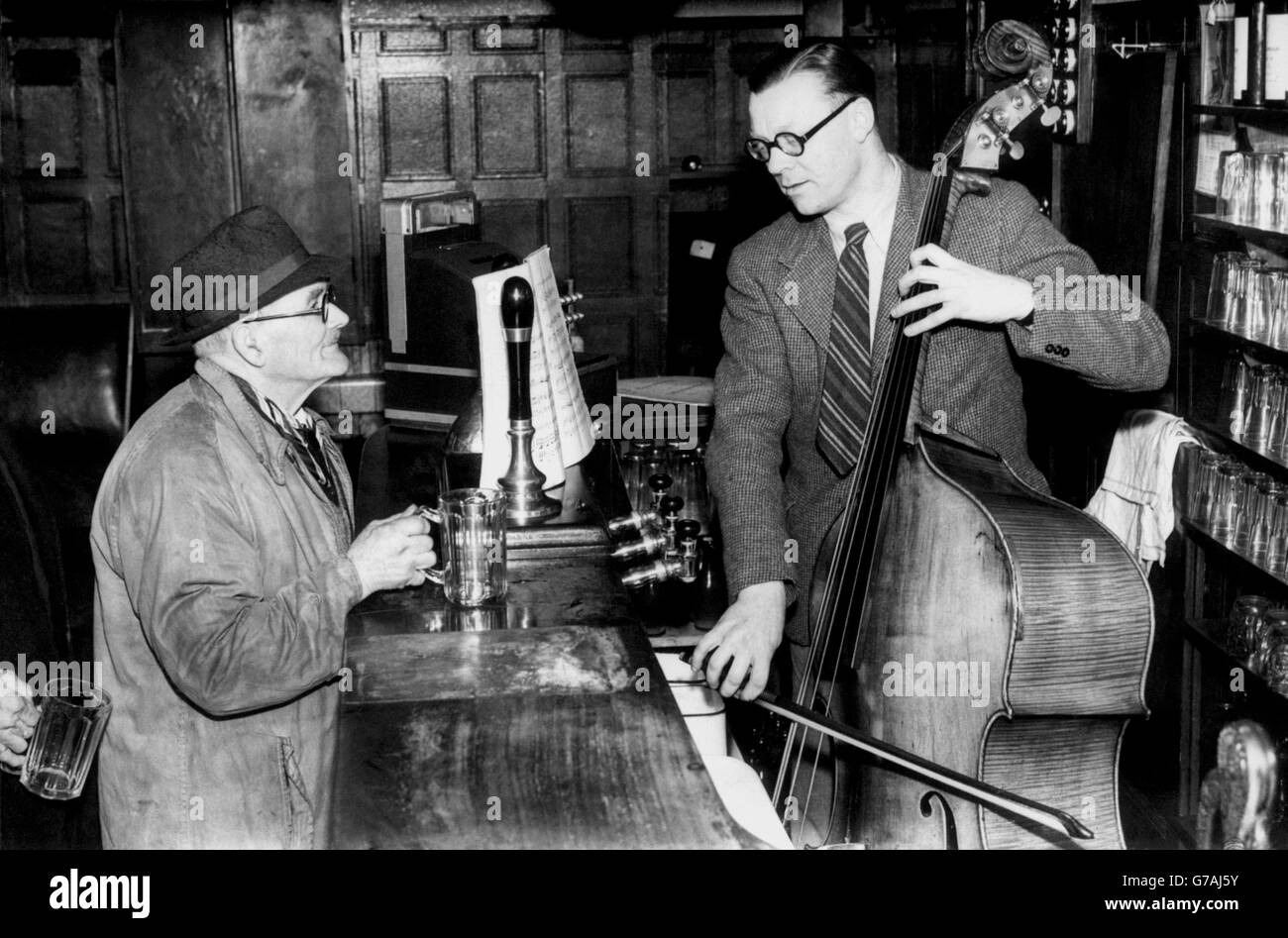 With his score propped against the beer pumps, Victor Cook, landlord of the Park-Lane Tavern, Cradley, near Wolverhampton, practices on his bull fiddle, while a thirsty customer waits with an empty glass. Stock Photo