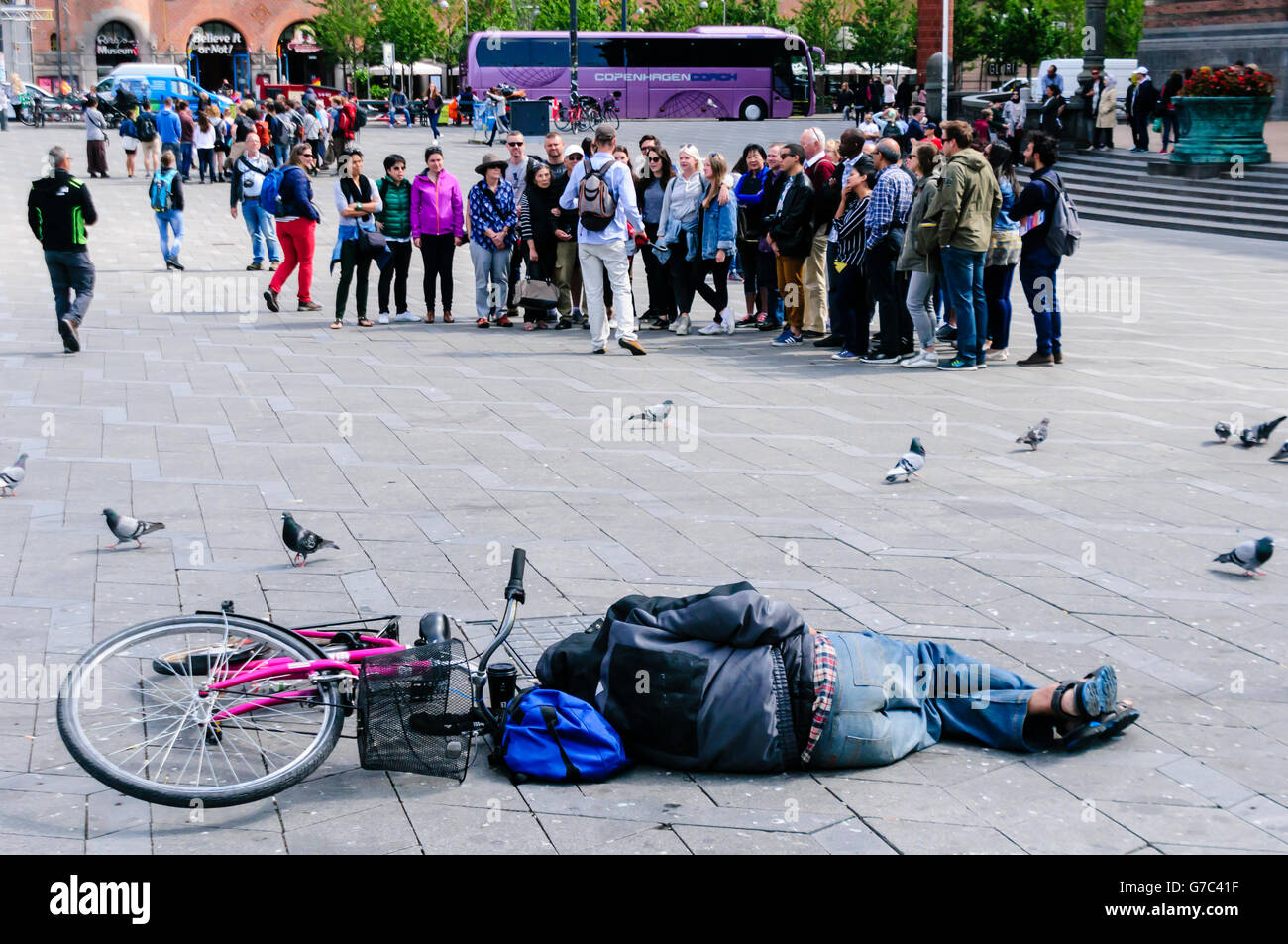 A drunk man sleeps in a public square beside his bicycle, as a group of tourists look on in Copenhagen, Denmark Stock Photo