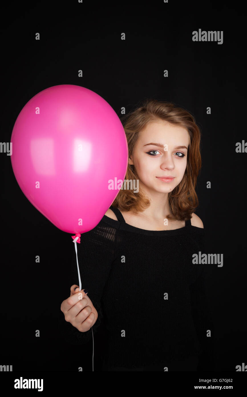 Studio portrait of smiling teenage Caucasian blond girl pointing with pink balloon over black background Stock Photo