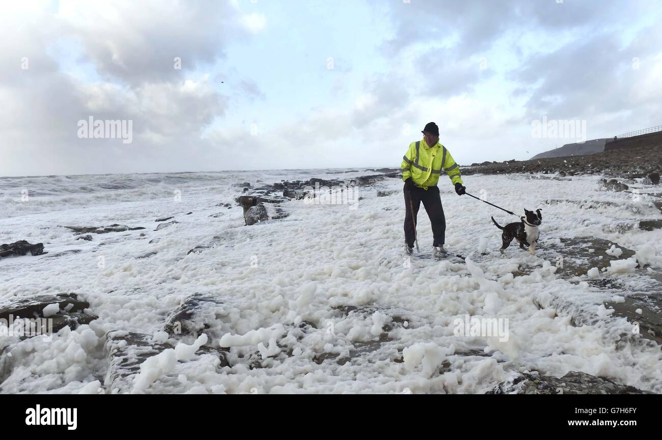A man and his dog walk through the foam blown from the rough sea near Whitehaven in Cumbria, as the stormy weather is causing disruption across parts of the UK with power cuts, ferry and train cancellations and difficult driving conditions. Stock Photo