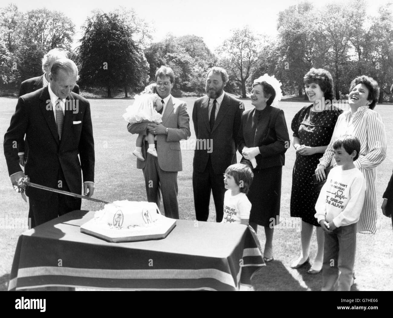 Patron of the British Heart Foundation, the Duke of Edinburgh in the grounds of Buckingham Palace cutting a heart-shaped cake to give a royal send-off to Heart Week '85. He is watched by Lord Soames (behind the Duke) and other former heart patients. The group, living proof of the Week's theme 'Research Saves Lives', are (l-r) Tania Didwell, 20 months and in the arms of her father, Peter Rawley, 51, Ann Tooth, 50, Lindsay Stickland, 30, Linda Rooney, 30, and at the front is Katherine Bradley, four, and Elliot Fleisher, eight. *Scanned low-res from print, high-res available on request* Stock Photo