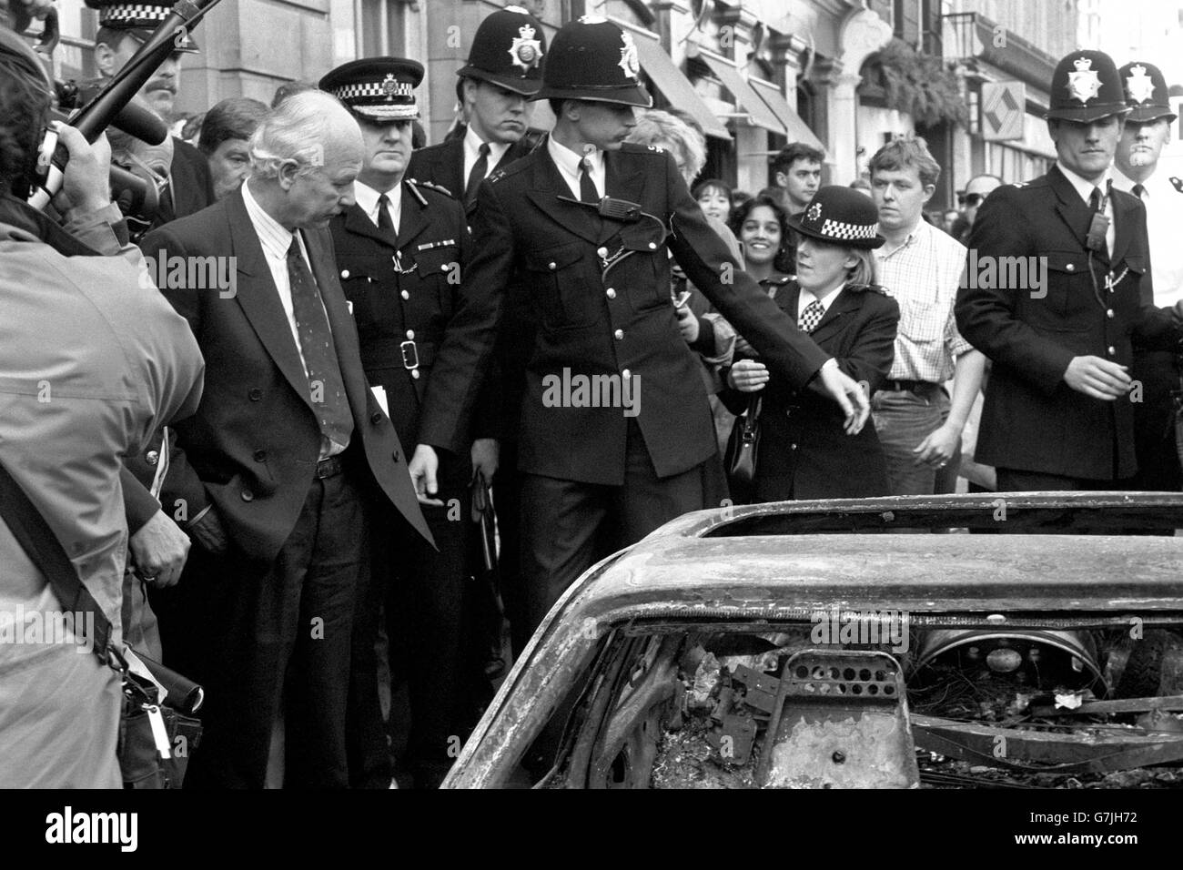 Police keep a heckling crowd at bay as Home Secretary David Waddington surveys the burnt-out wreck of a car in central London, following the poll tax riots yesterday. Stock Photo