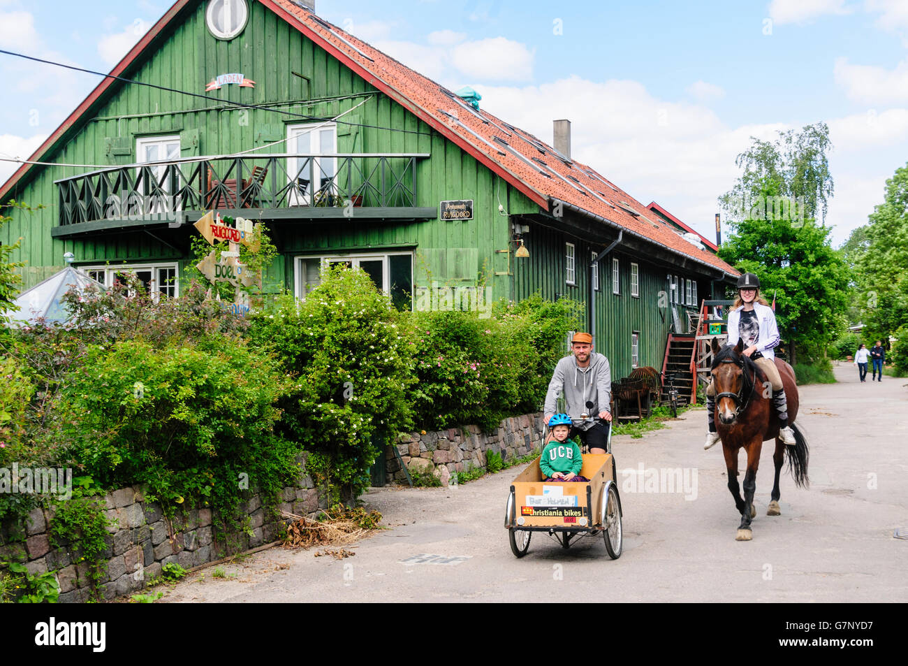 People a bicycle and horse pass a traditional green wooden house with terracotta tiled roof in Freetown Christiania, Copenhagen Stock Photo