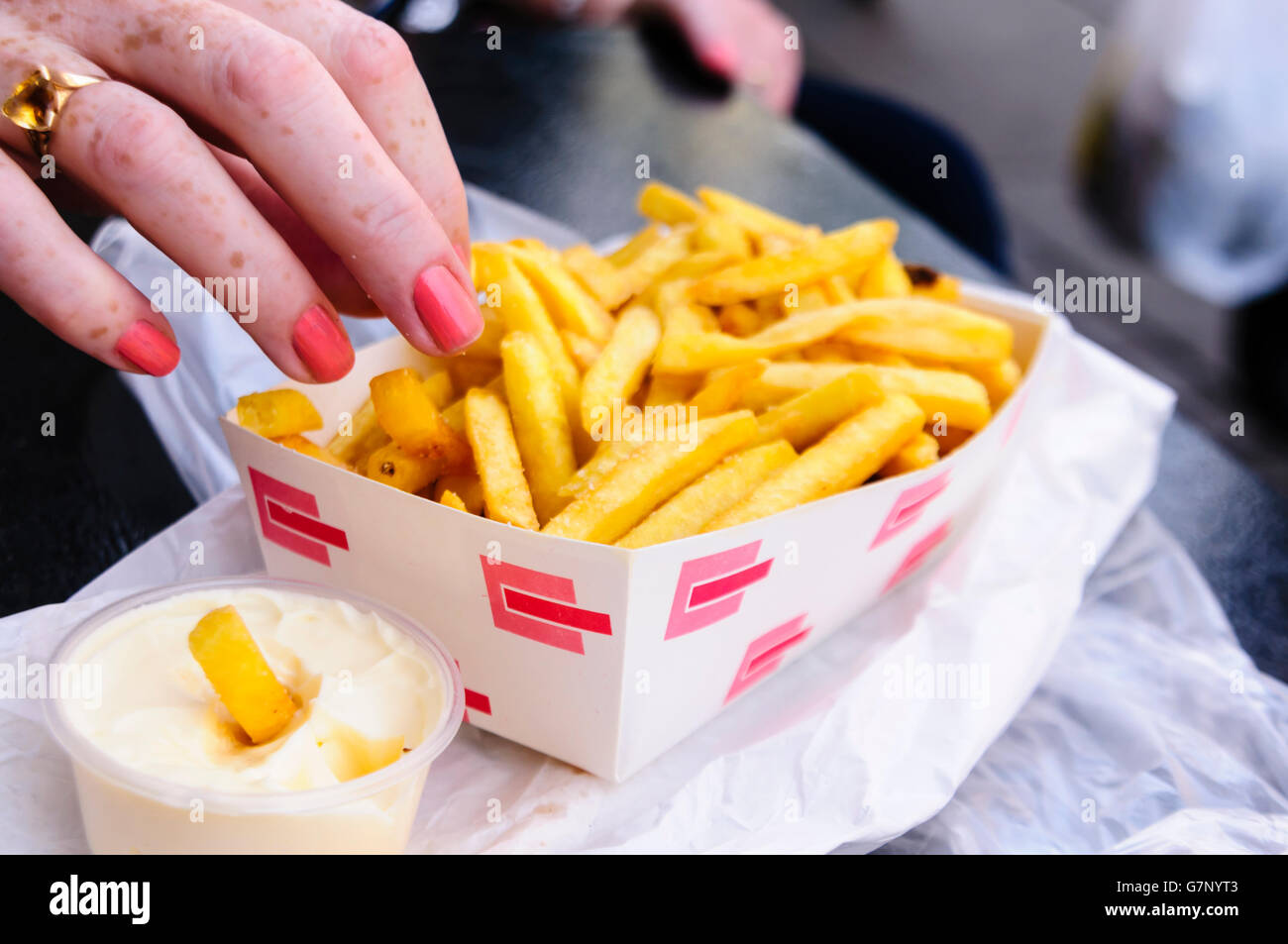 A woman takes a fry from a box of French Fries beside a tub of mayonnaise. Stock Photo