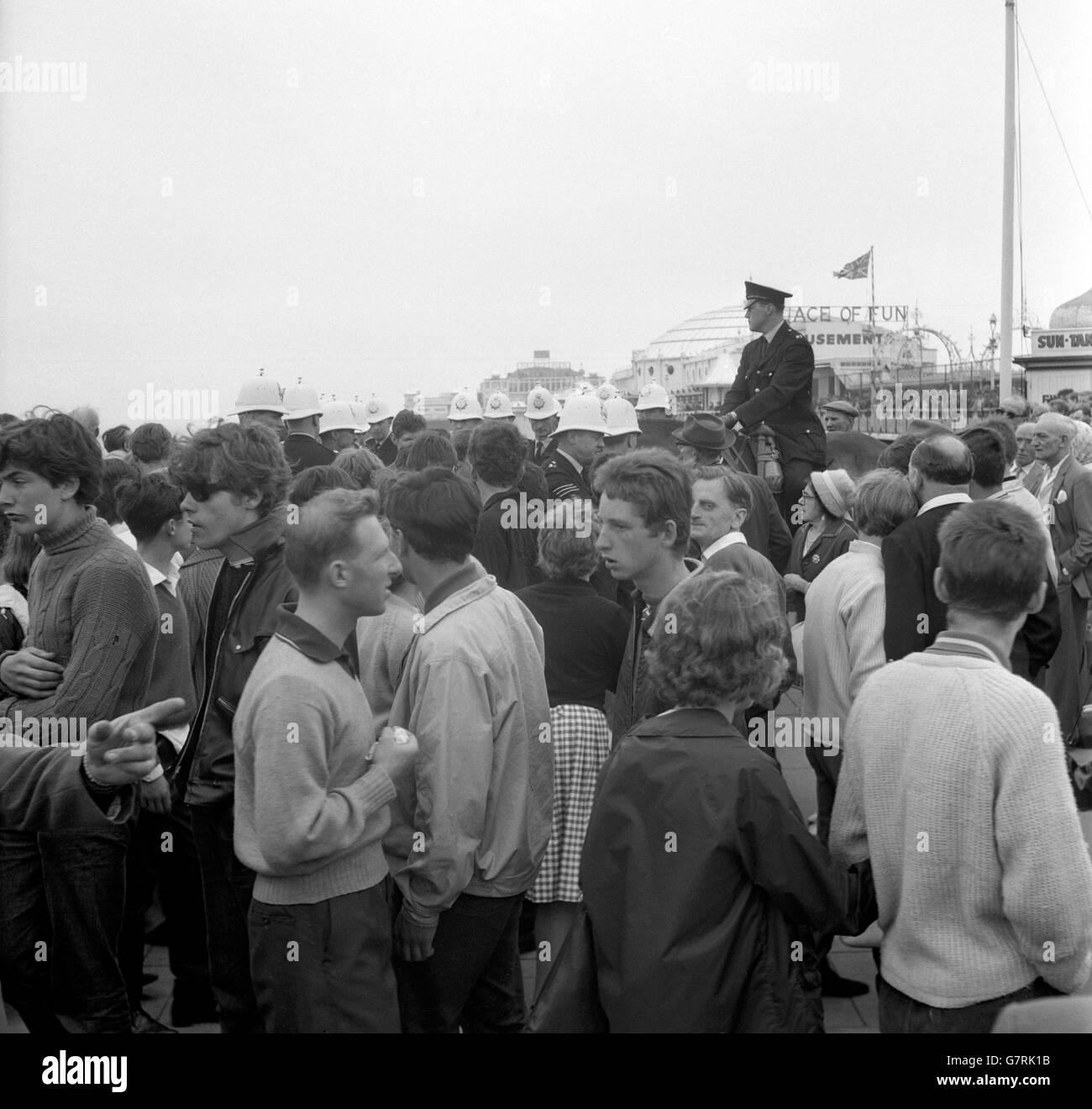 A mounted policeman and white helmeted colleagues move among the crowd on the seafront at Brighton, Sussex, where trouble flared between rival gangs of Mods and Rockers. There was a sharp battle between rival gangs on the steps of the Aquarium, when deckchairs and rubbish were thrown. Some of the rockers escaped by dropping 15ft over a wall to the promenade below. There were also pitched battles at Margate, Kent, where two youths were taken to hospital with stab wounds. Stock Photo