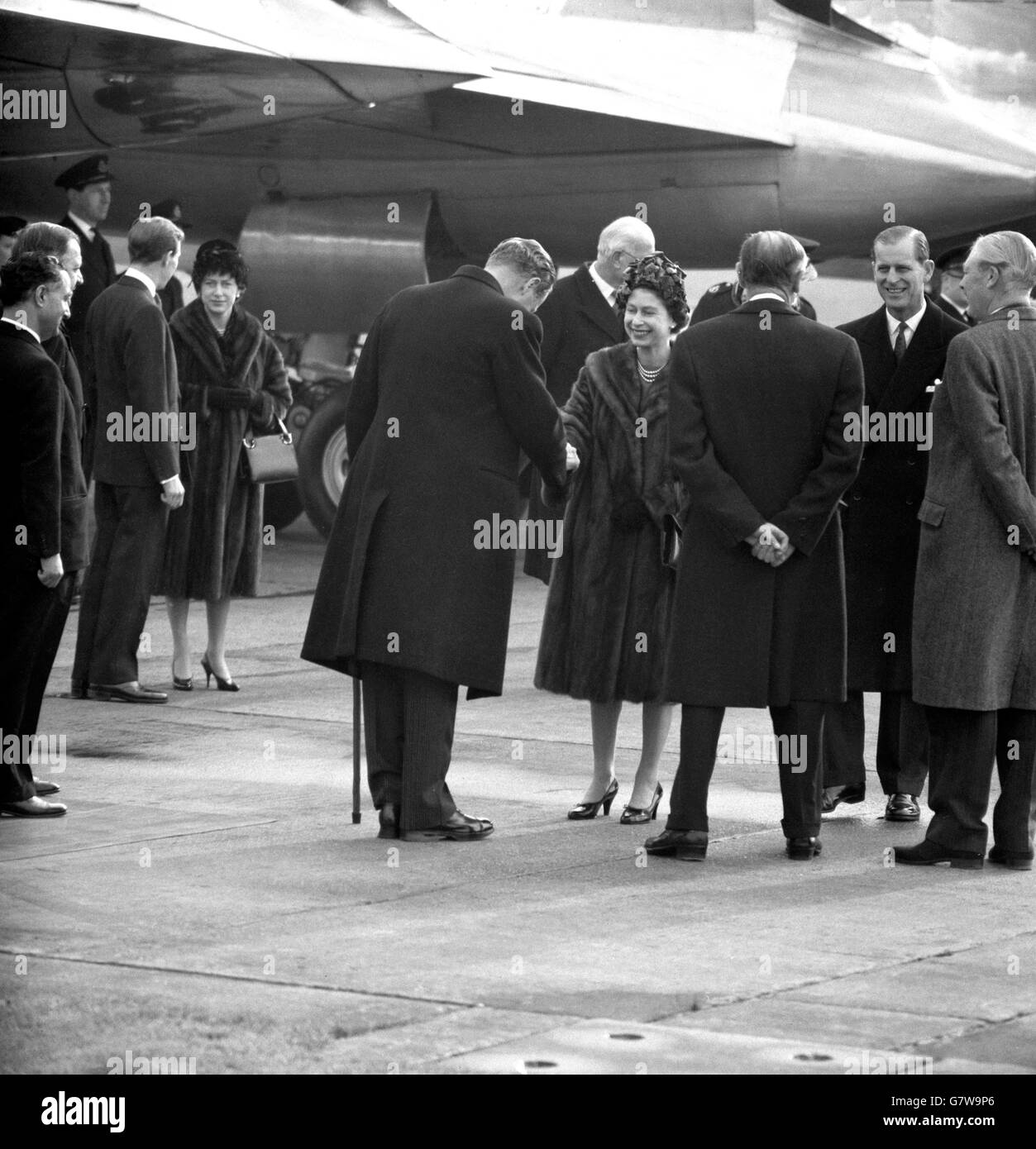 Smiling, Queen Elizabeth II and Prince Philip the Duke of Edinburgh say goodbye with handshakes at London Airport before boarding a BOAC Britannia liner to fly to New Delhi. Far right is the Prime Minister, Harold Macmillan. The Queen, wearing a light brown mink coat over a royal blue dress and a hat of royal blue and vivid green velvet petals, and the Duke were leaving for a 20,000 mile tour of India, Pakistan, Nepal and Iran. Stock Photo