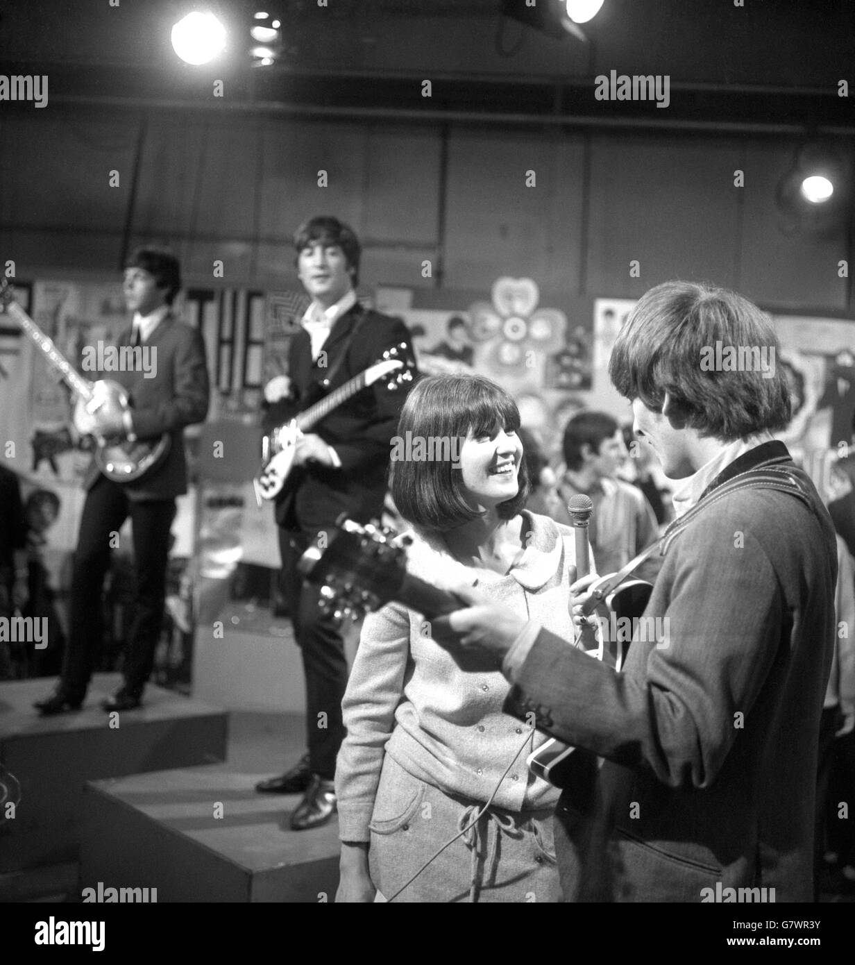 The crowds of Beatles fans outside the building had had to struggle gust to get a glimpse of their idols. But here's a girl inside Television House, Kingsway, London, who could actually talk to them - officially. She is CATHY McGOWAN, teenage adviser to the Independent television pop programme 'Ready, Steady, Go', and is seen interviewing GEORGE HARRISON at the rehearsal which preceded The Beatles' appearance. The two members of the group seen in background are PAUL McCARTNEY (left) and JOHN LENNON. Stock Photo