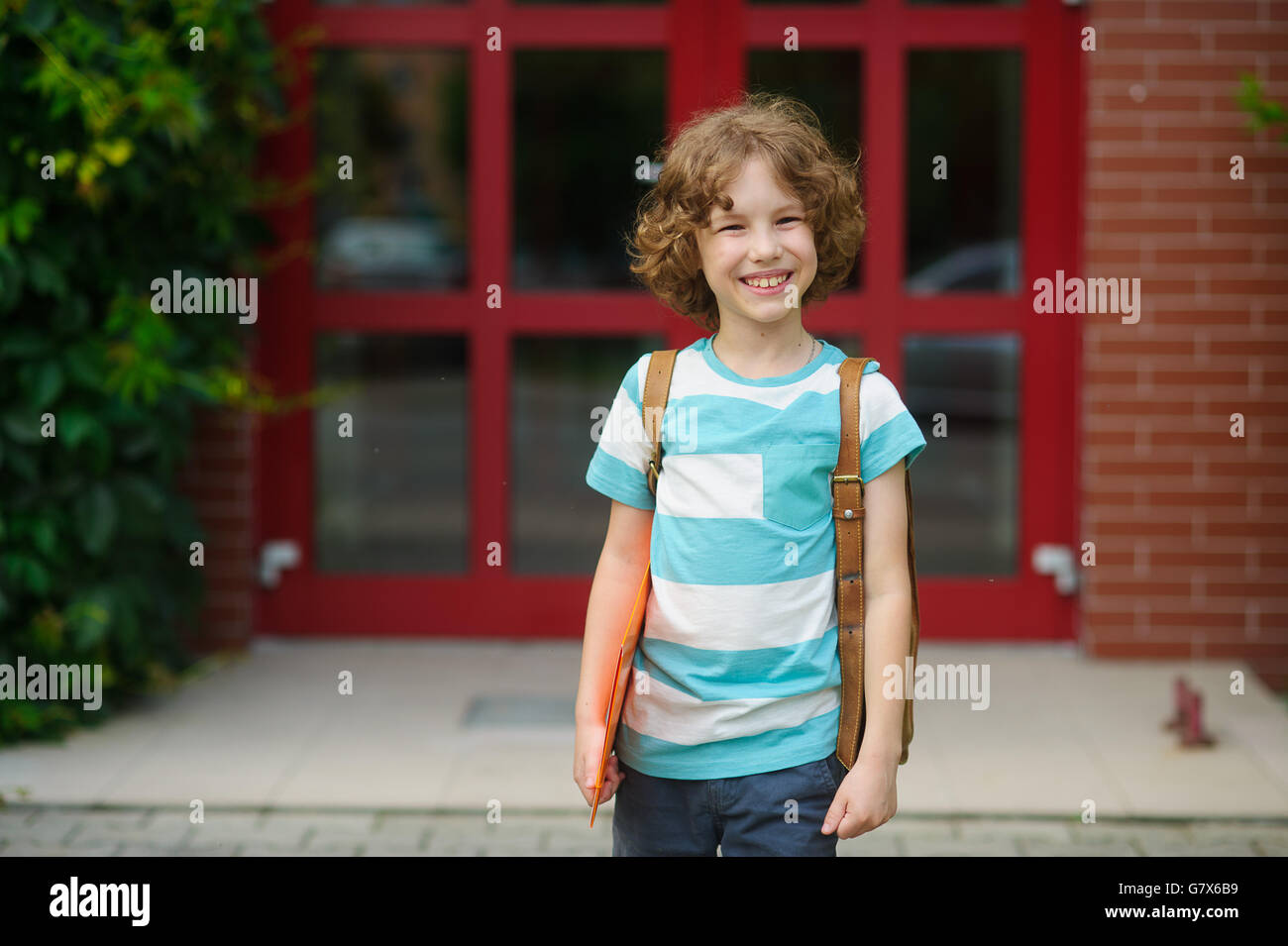The little schoolboy stand on a schoolyard and joyfully smiles. The curly boy with a nice face. Behind the school student's shou Stock Photo