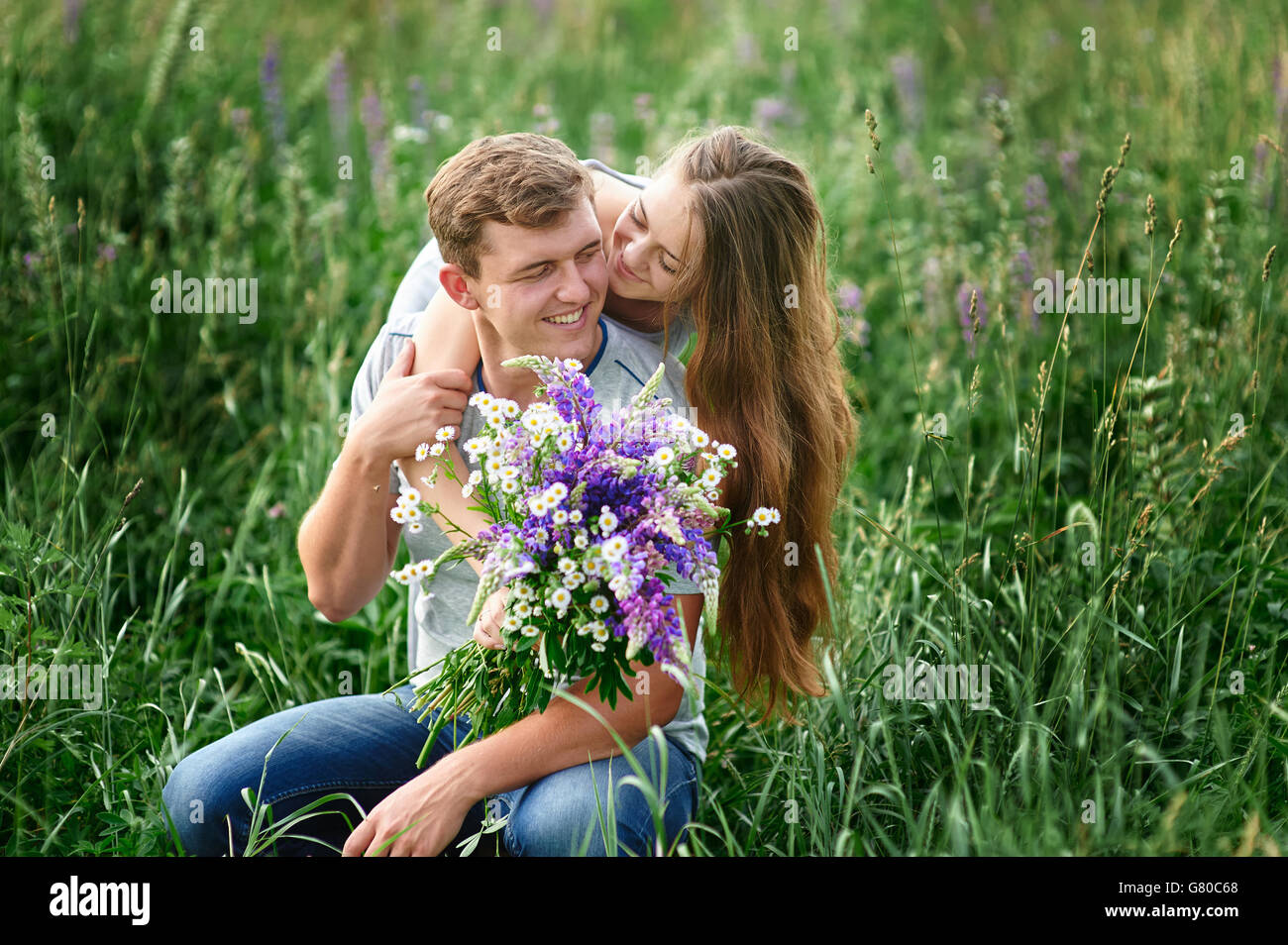 beautiful woman with bouquet of wild flowers hugging man sitting on the grass in meadow Stock Photo