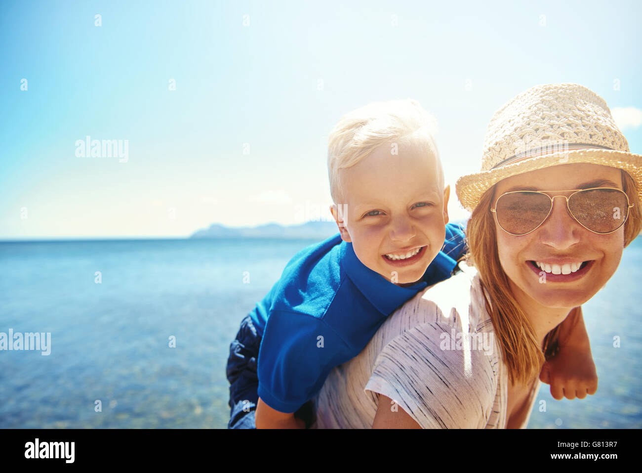 Happy little boy getting a piggy back ride from his attractive smiling mother in sunhat and sunglasses as they relax on a tropic Stock Photo
