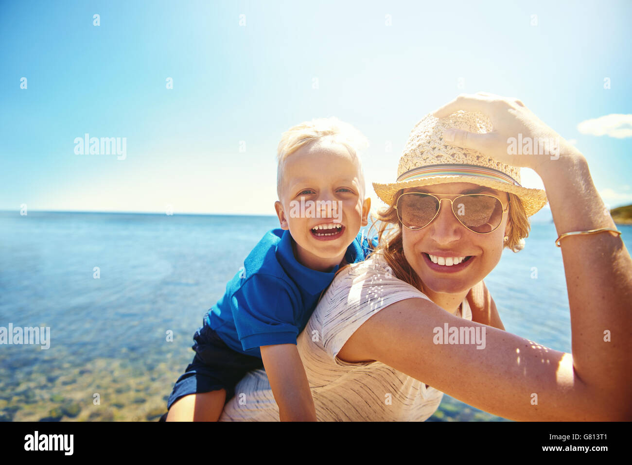 Happy young mother and son on a tropical beach with the laughing little boy getting a piggy back ride on her back Stock Photo