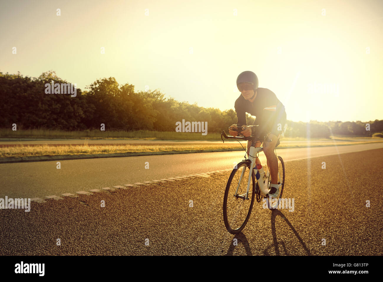 Single male bicyclist wearing shorts, helmet and sunglasses training for long distance racing on road bike at sunrise with copy Stock Photo