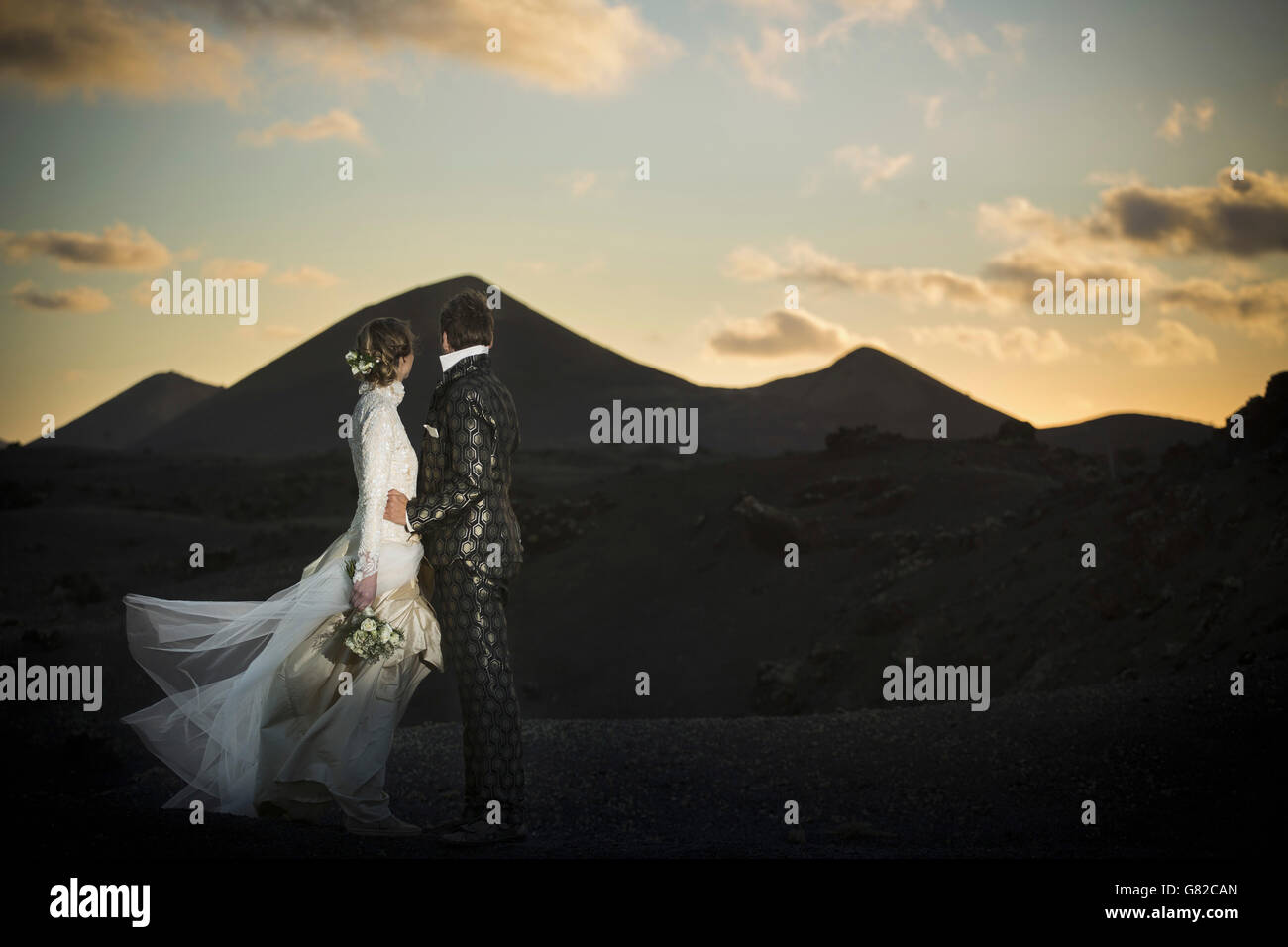 Full length side view of loving bridge and groom standing on volcanic landscape during sunset Stock Photo