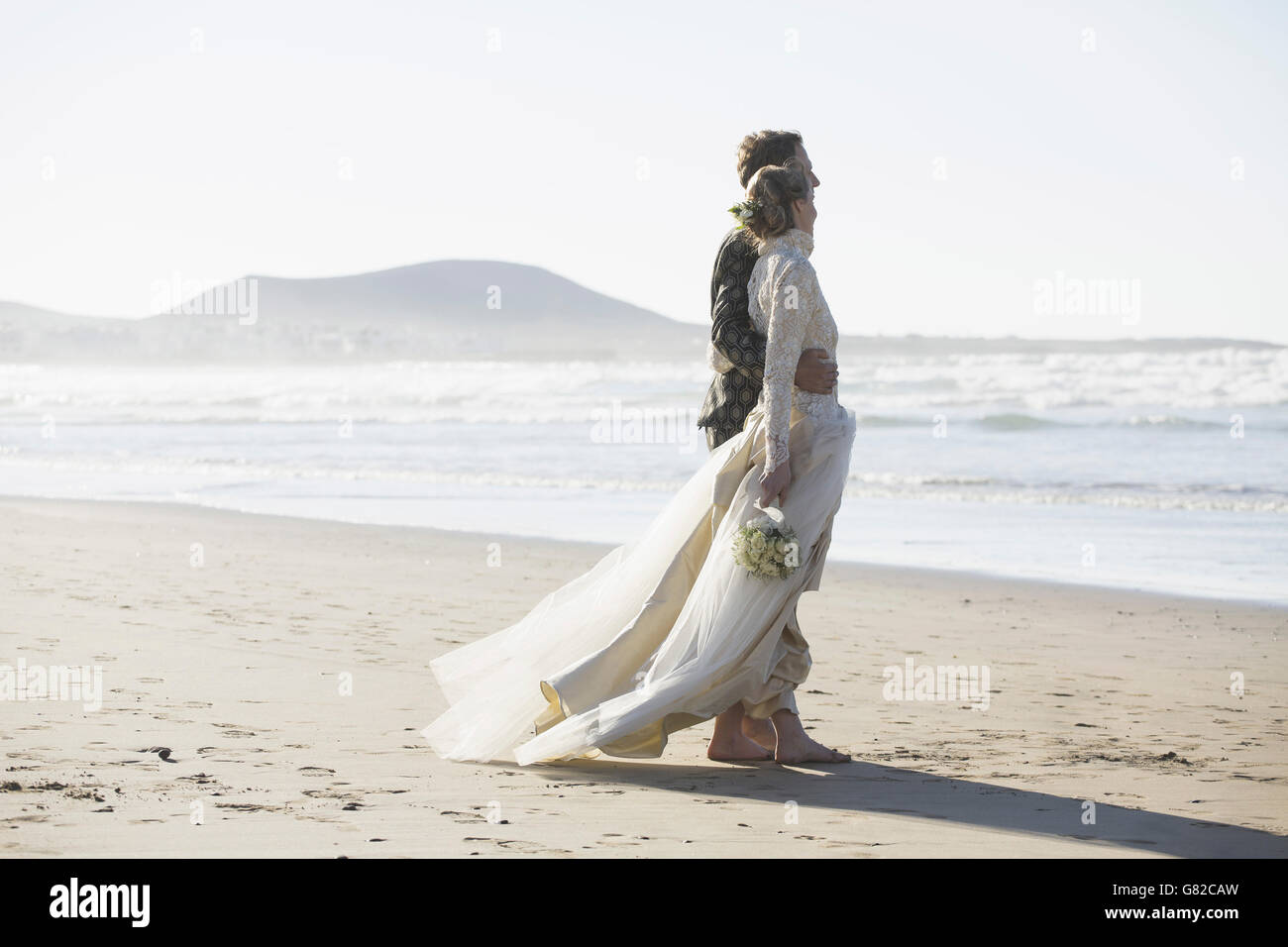 Full length side view of bride and groom standing arm around at beach Stock Photo