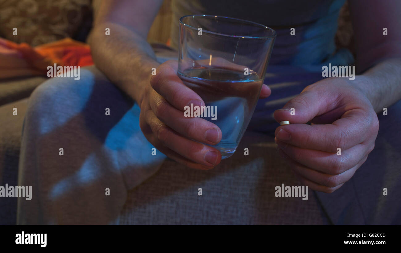 Midsection of holding painkiller and drinking glass while sitting on sofa Stock Photo