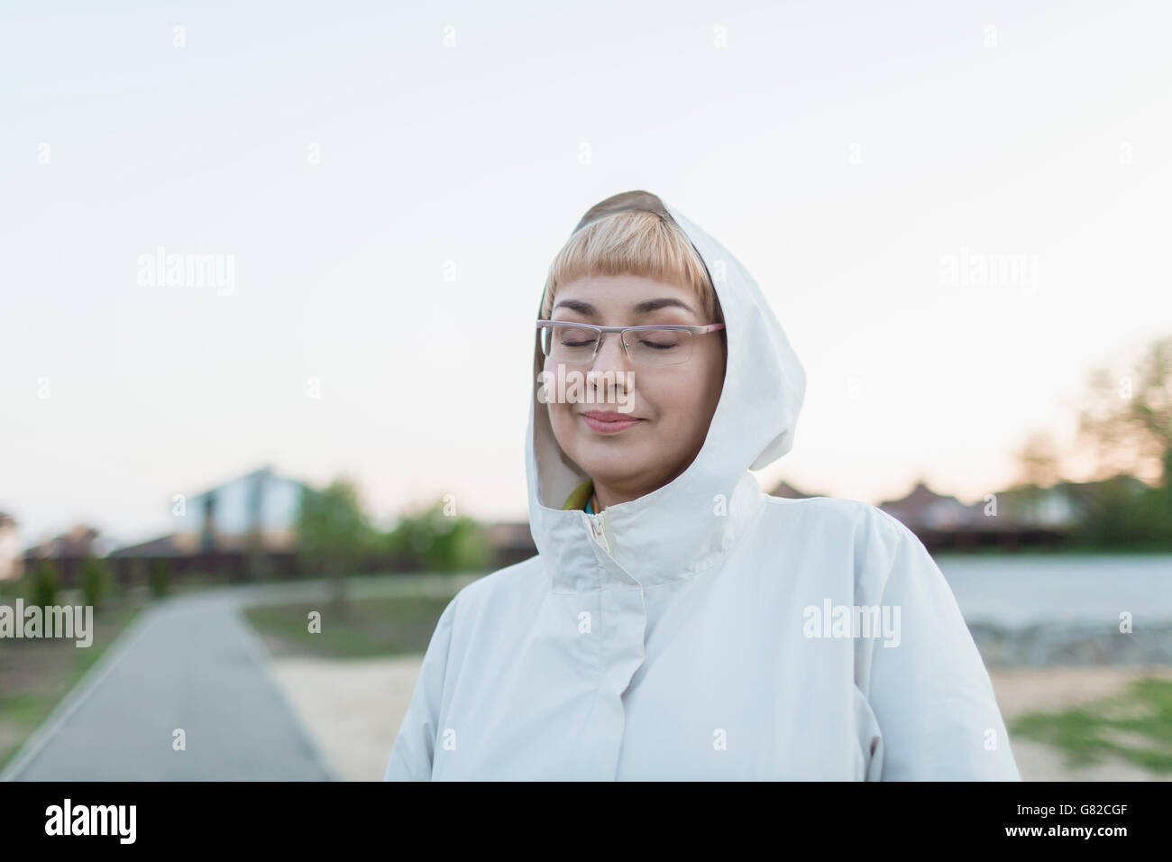 Smiling woman with closed eyes wearing hood in park against clear sky Stock Photo