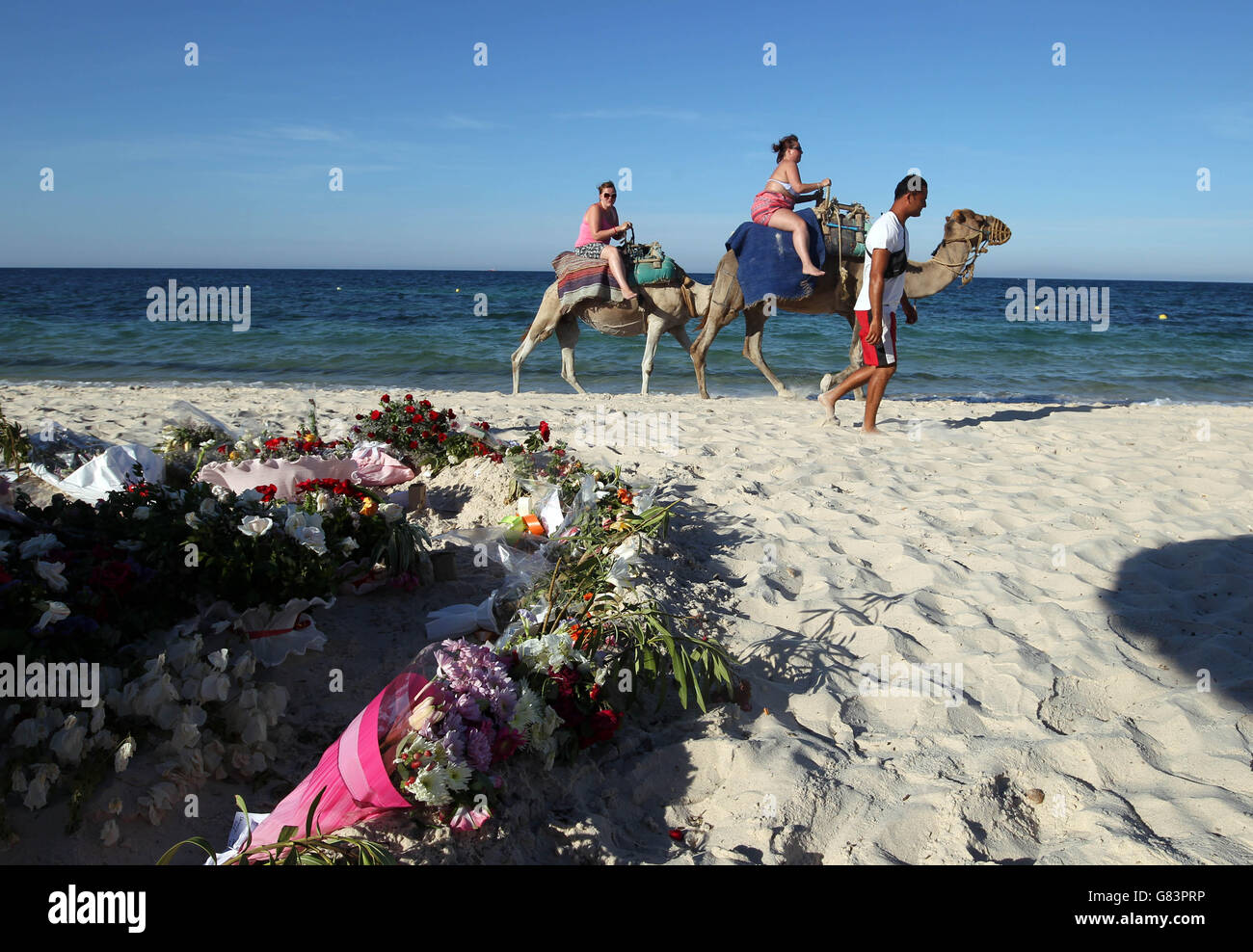 People on camels pass tributes on the beach near the RIU Imperial Marhaba hotel in Sousse, Tunisia, as British holidaymakers defy the terrorists and continue to stay in Sousse despite the bloodbath on the beach. PRESS ASSOCIATION Photo. Picture date: Tuesday June 30, 2015. The sands at Sousse were quiet and calm today as tourists and locals alike continued to pay their respects to the 38 dead outside the RIU Imperial Marhaba and Bellevue hotels. Flowers continue to be laid at three heart-shaped memorials that mark where so many people lost their lives, with many people in tears as they read Stock Photo