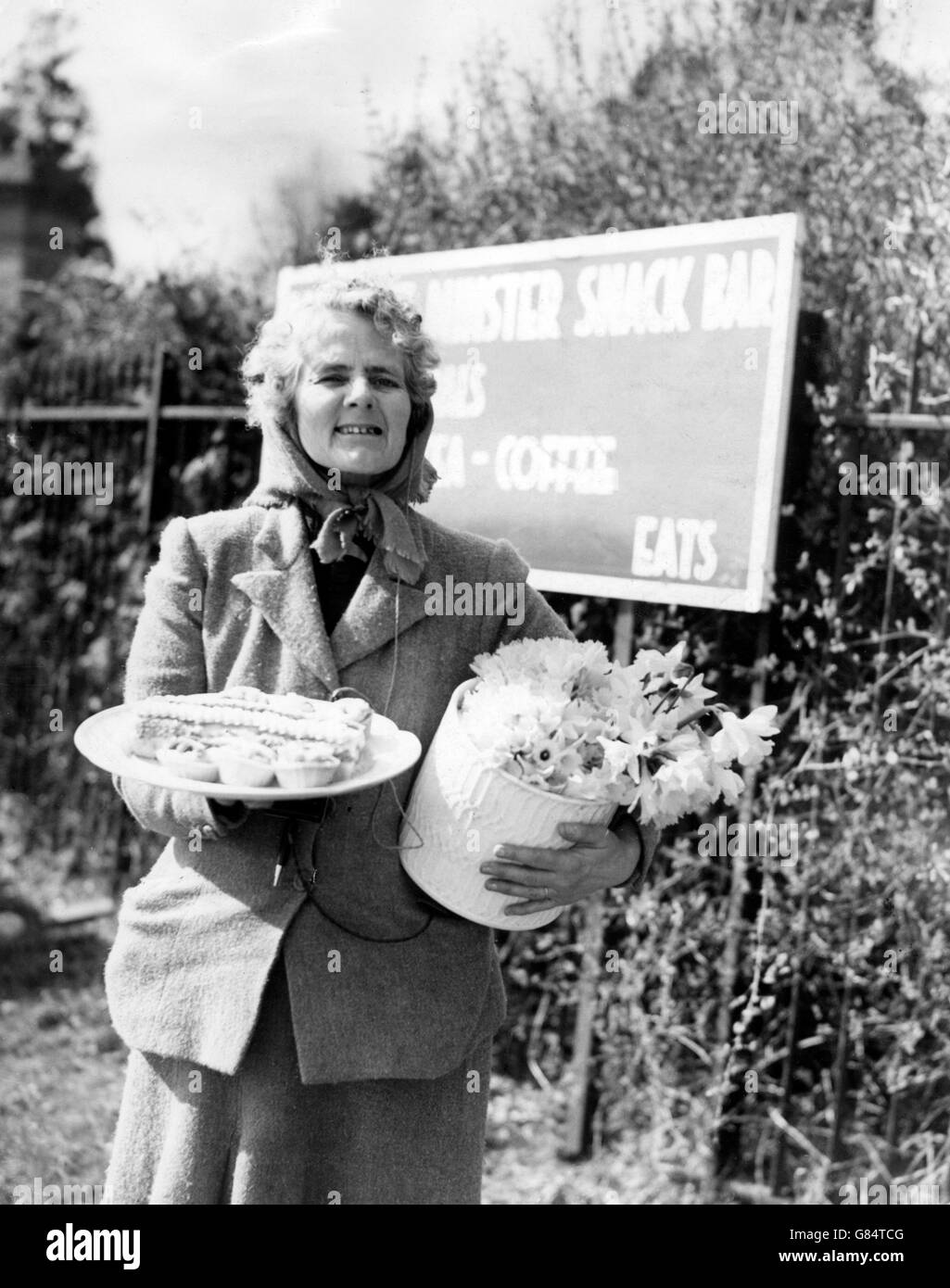 Holding a plate of home-baked cakes and a jar of daffodils is Lady Lees, wife of the third baronet, 54-year-old Sir John Victor Elliott Lees. She is in front of a sign advertising her new snack bar at the gates of the Manor House, Lytchett Minster, at Poole in Dorset. Stock Photo