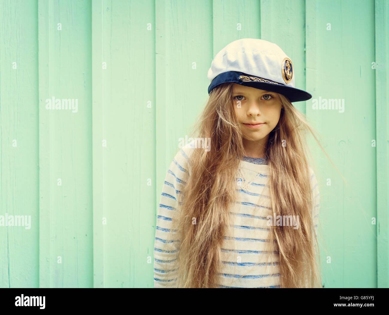 Cute little girl stands near a turquoise wall in sailor hat and smiling Space for text. Stock Photo