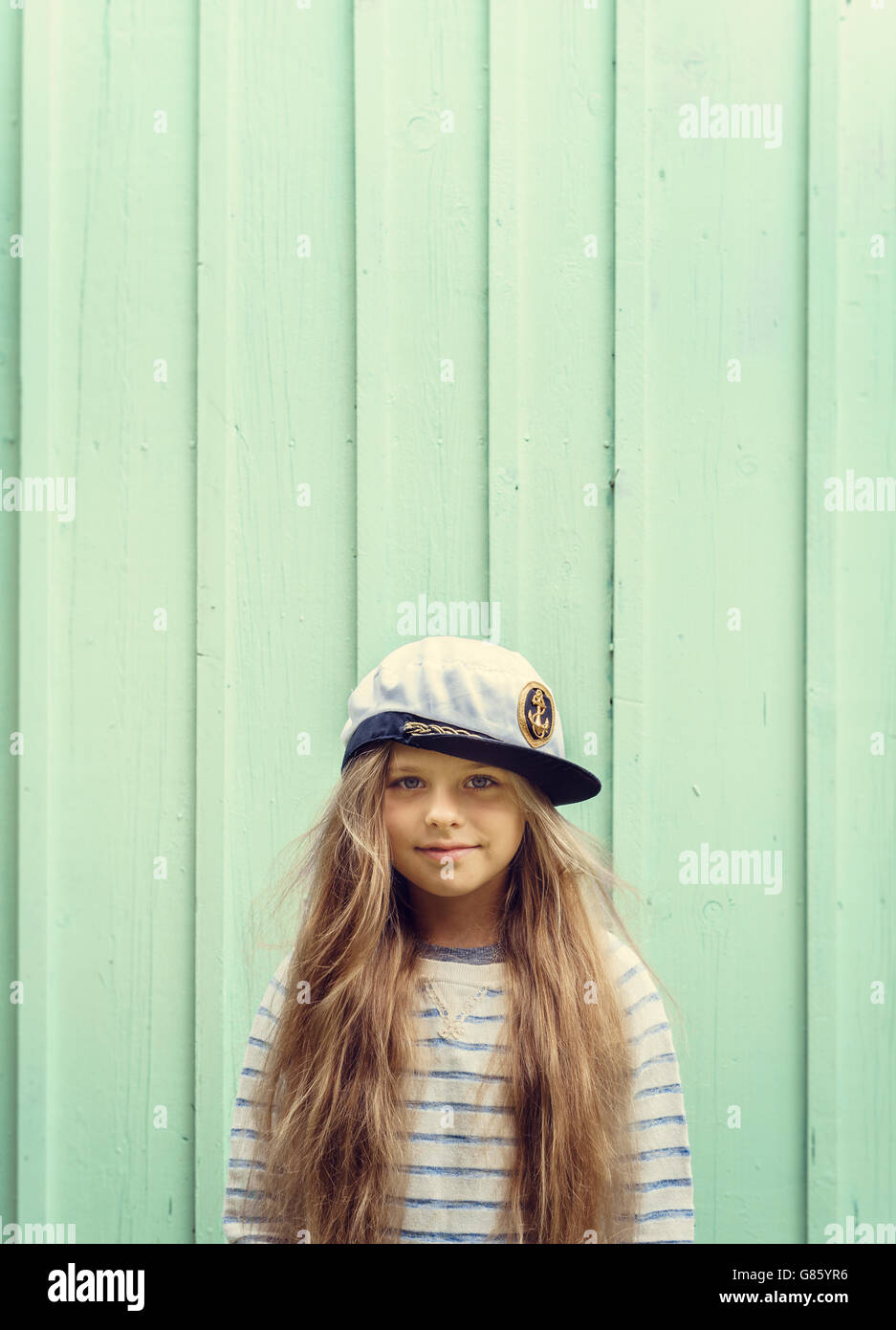 Cute little girl stands near a turquoise wall in sailor hat and smiling Space for text. Stock Photo